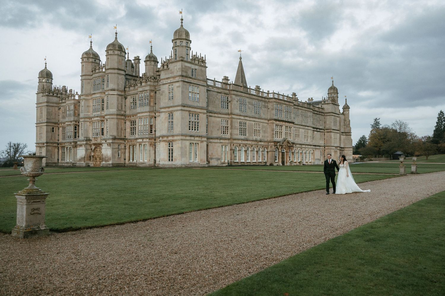 Bride and groom stand before the grand Burghley House estate on their wedding day, surrounded by manicured lawns.