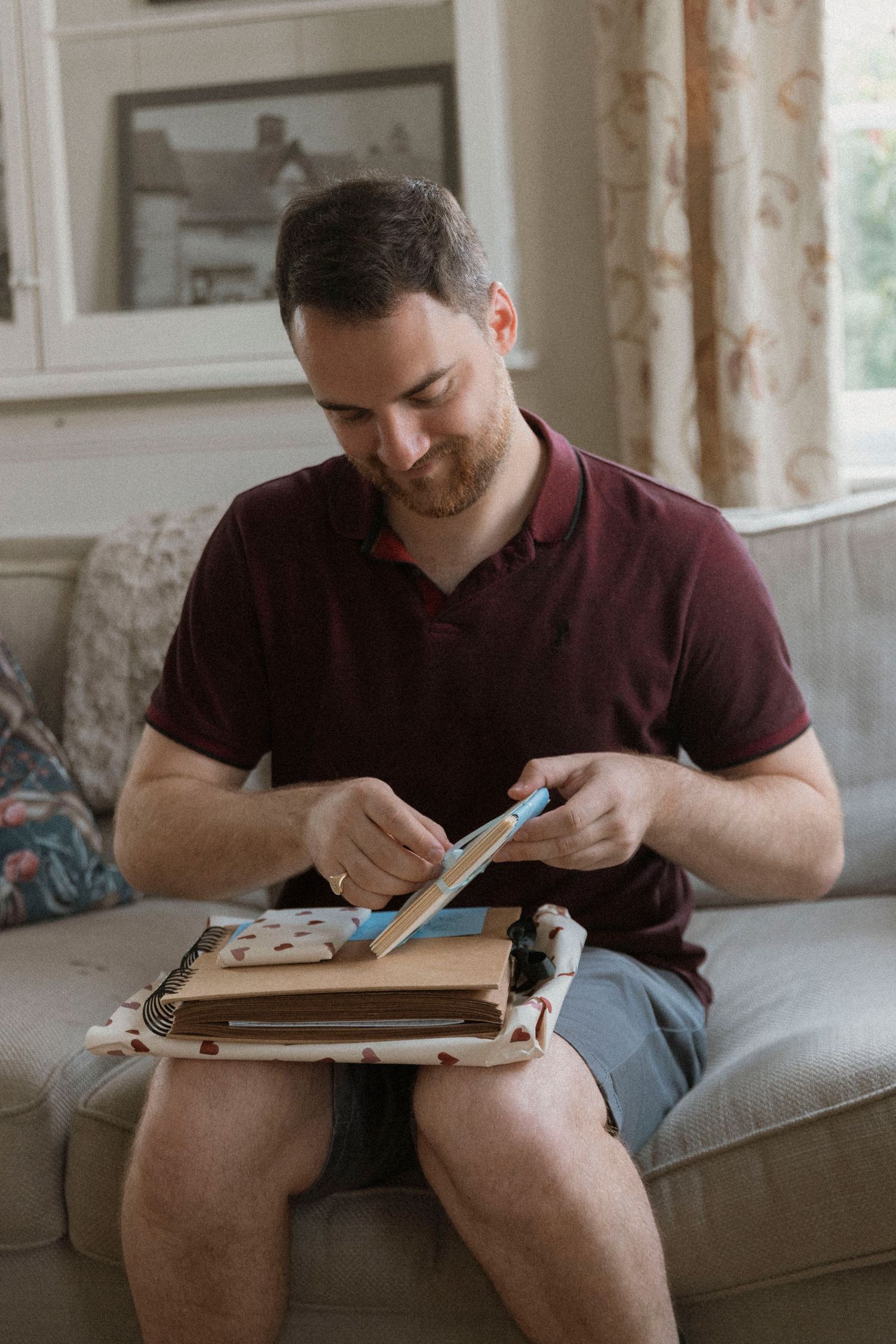 Smiling man in maroon polo shirt sitting on sofa, writing in a wooden clipboard journal with a blue pen.
