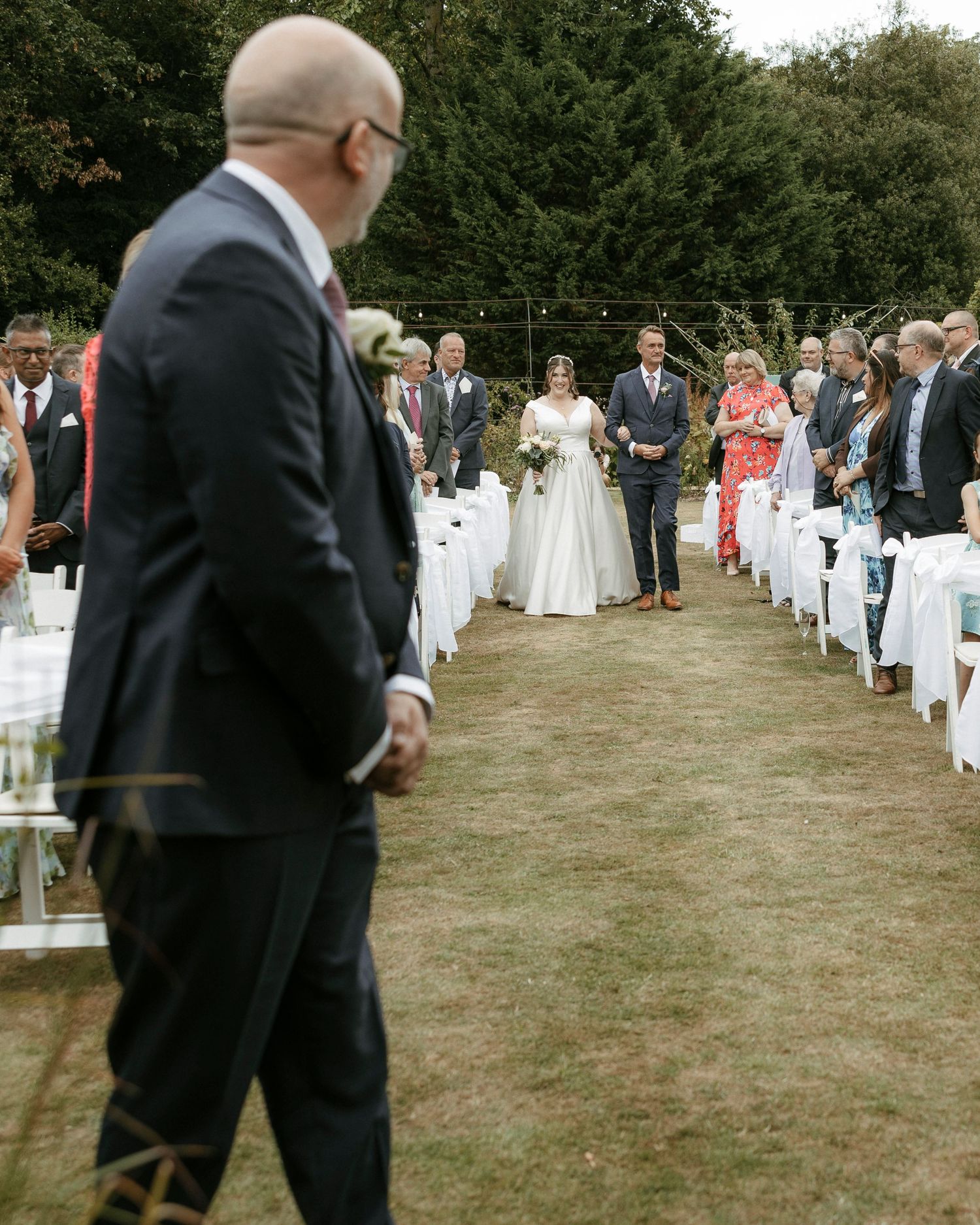 Bride walks down outdoor wedding aisle in white gown toward groom, guests seated on white chairs in garden ceremony.