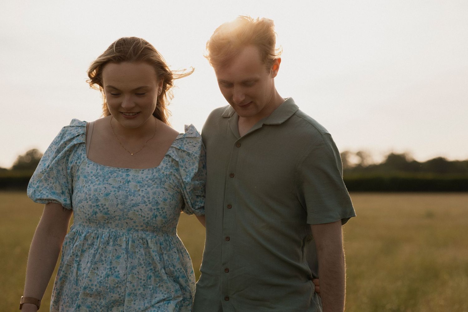 Couple walking hand in hand through a golden field at sunset, smiling warmly.