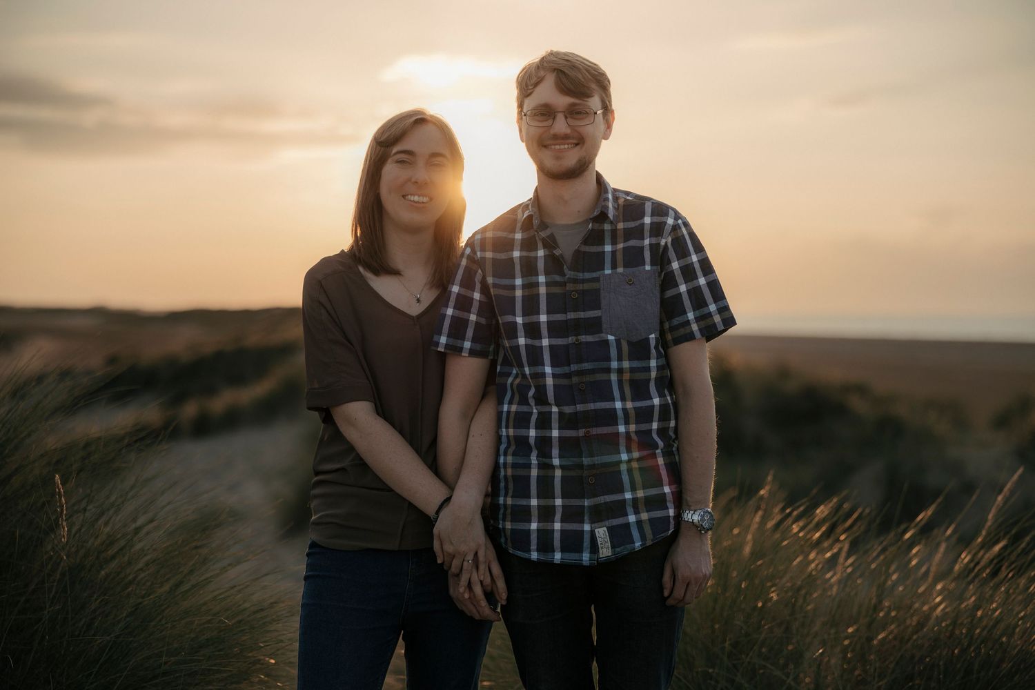 Couple holding hands at golden hour sunset in grassy dunes, romantic outdoor portrait photography.