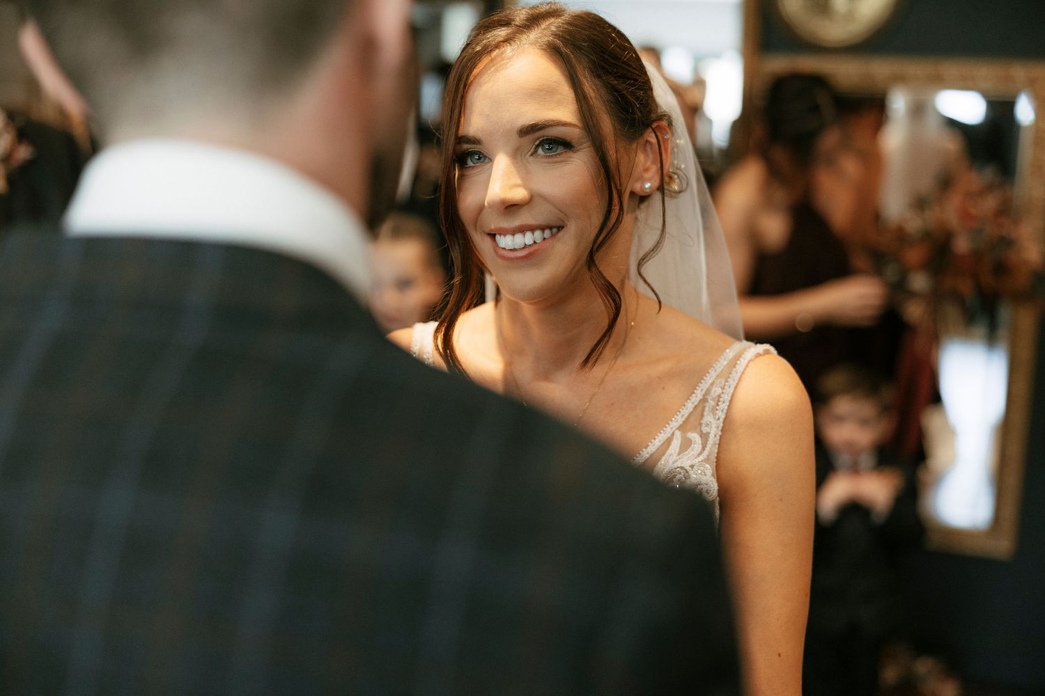 Smiling bride in lace gown and veil during wedding ceremony, exchanging vows with groom in plaid suit.