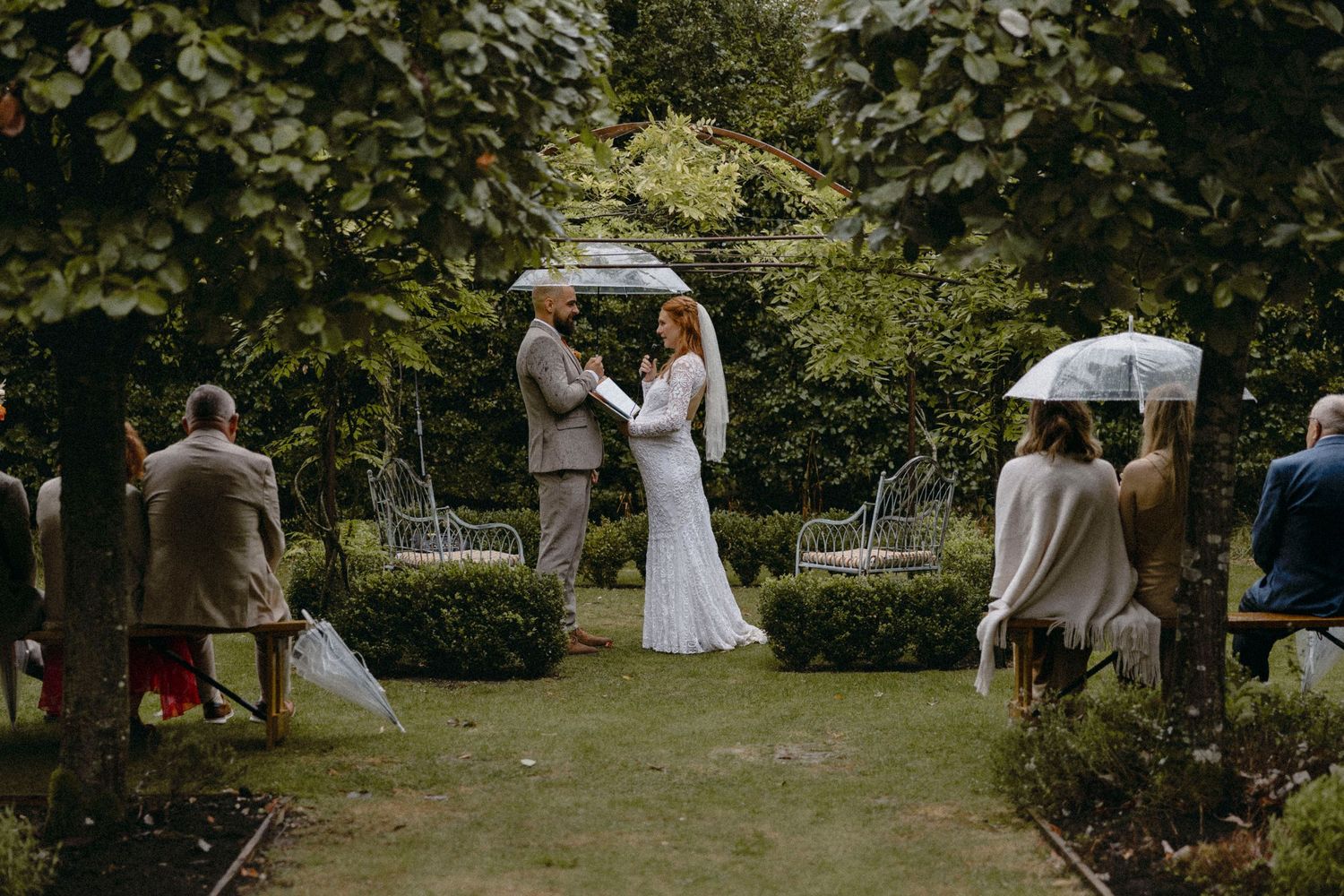 Couple exchanging vows in a lush garden wedding ceremony with guests holding umbrellas on a cloudy day.
