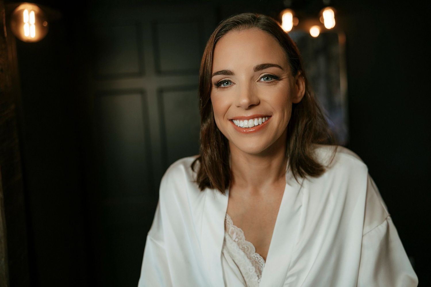A person in a white robe smiles warmly at the camera in a dimly lit room with glowing sconce lights.