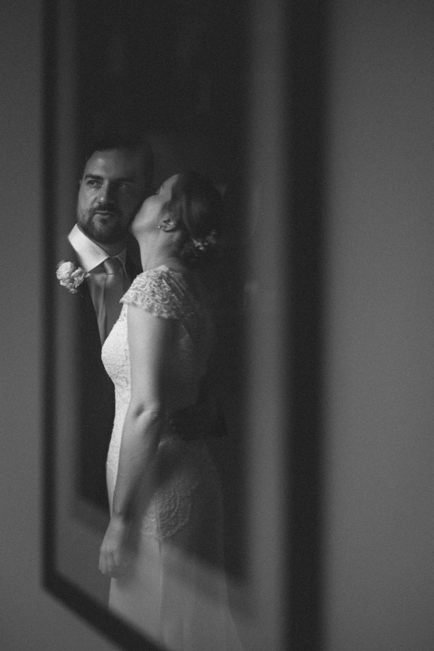 Black and white wedding photo of bride and groom sharing an intimate moment, captured through a doorway.