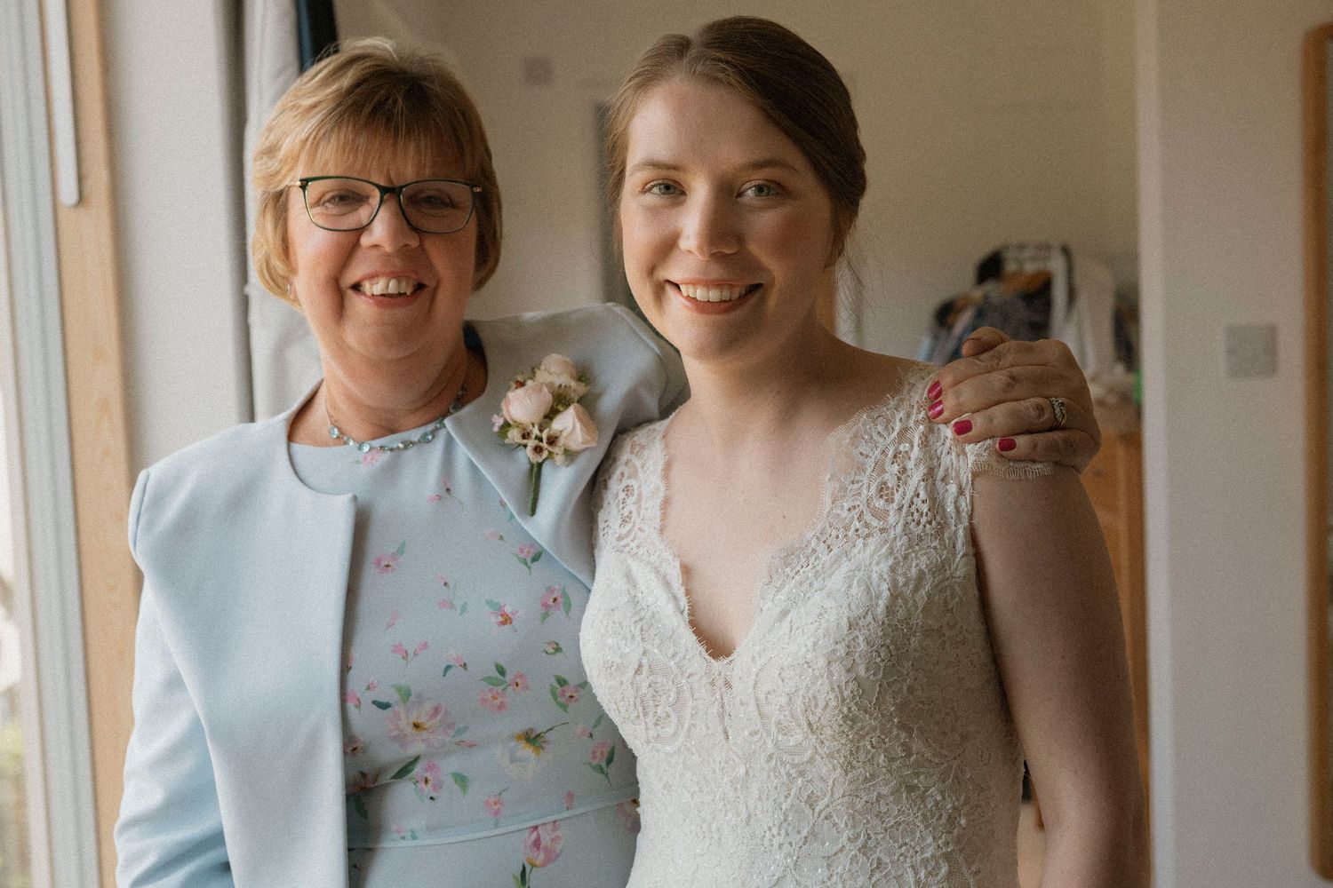 Bride in lace wedding dress smiles with older woman in blue floral outfit, both posing happily indoors.