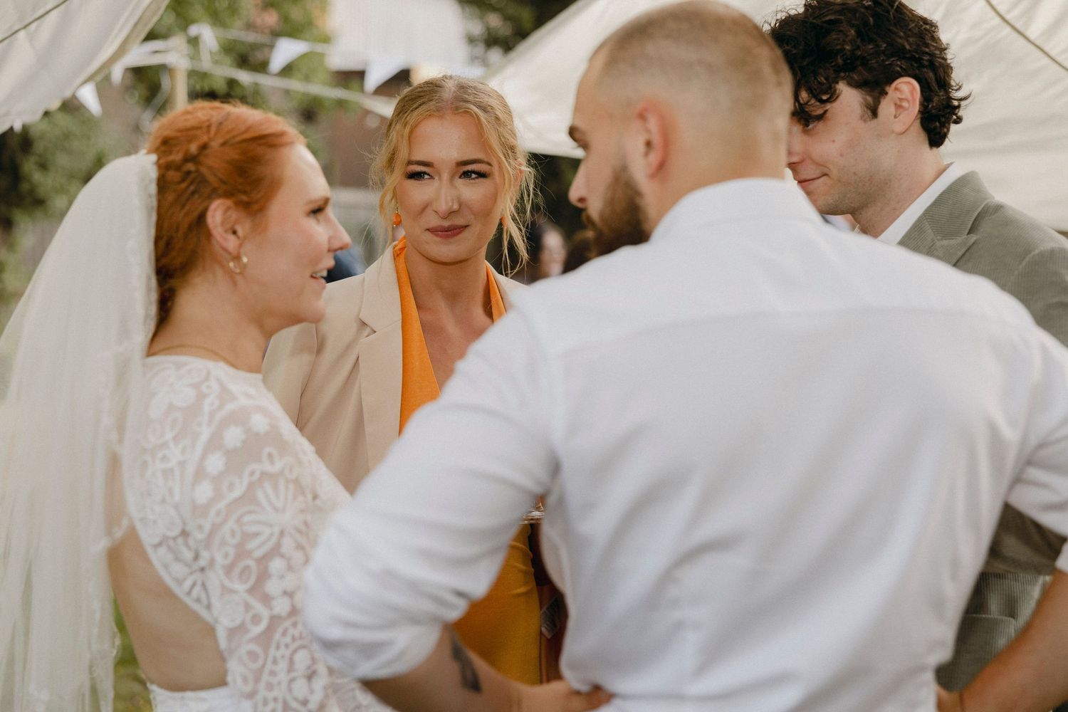 Bride and groom exchanging vows with officiant and guest at an outdoor wedding ceremony under a white tent.