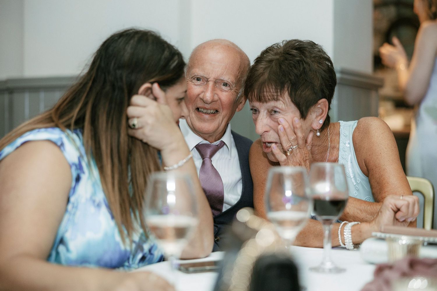 Three people sharing a warm moment at a dining table with wine glasses, whispering and smiling at a formal event.