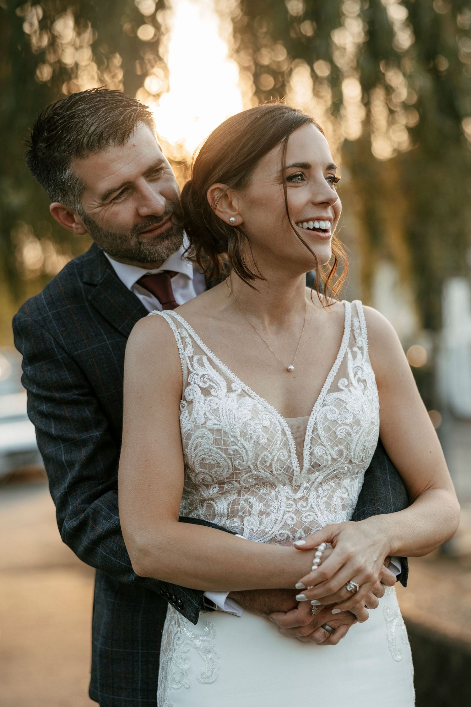 A joyful bride in a lace gown laughs as her bearded groom in a plaid suit embraces her during golden hour.