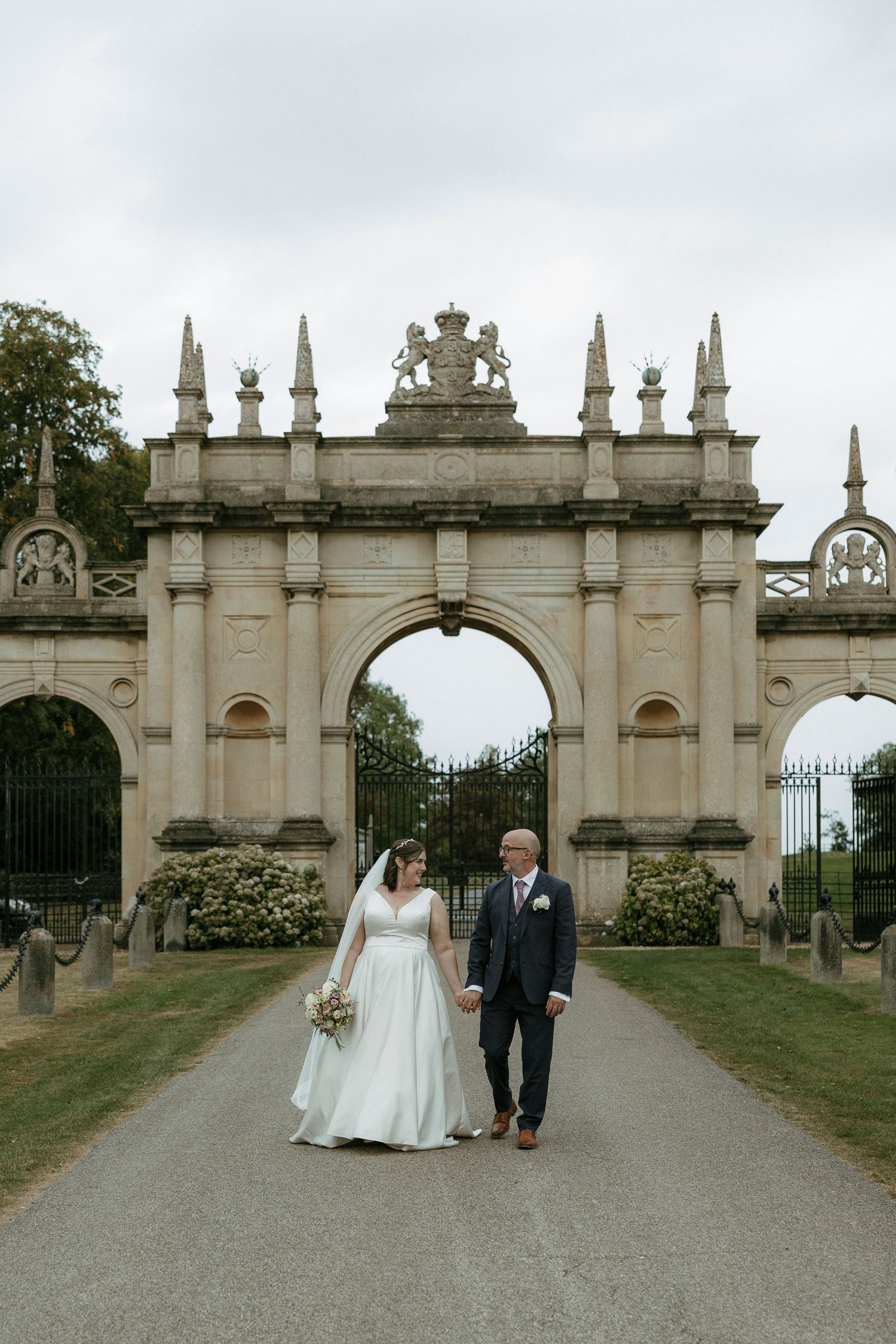 Bride and groom hold hands walking toward ornate stone arch gate at elegant English estate wedding venue.