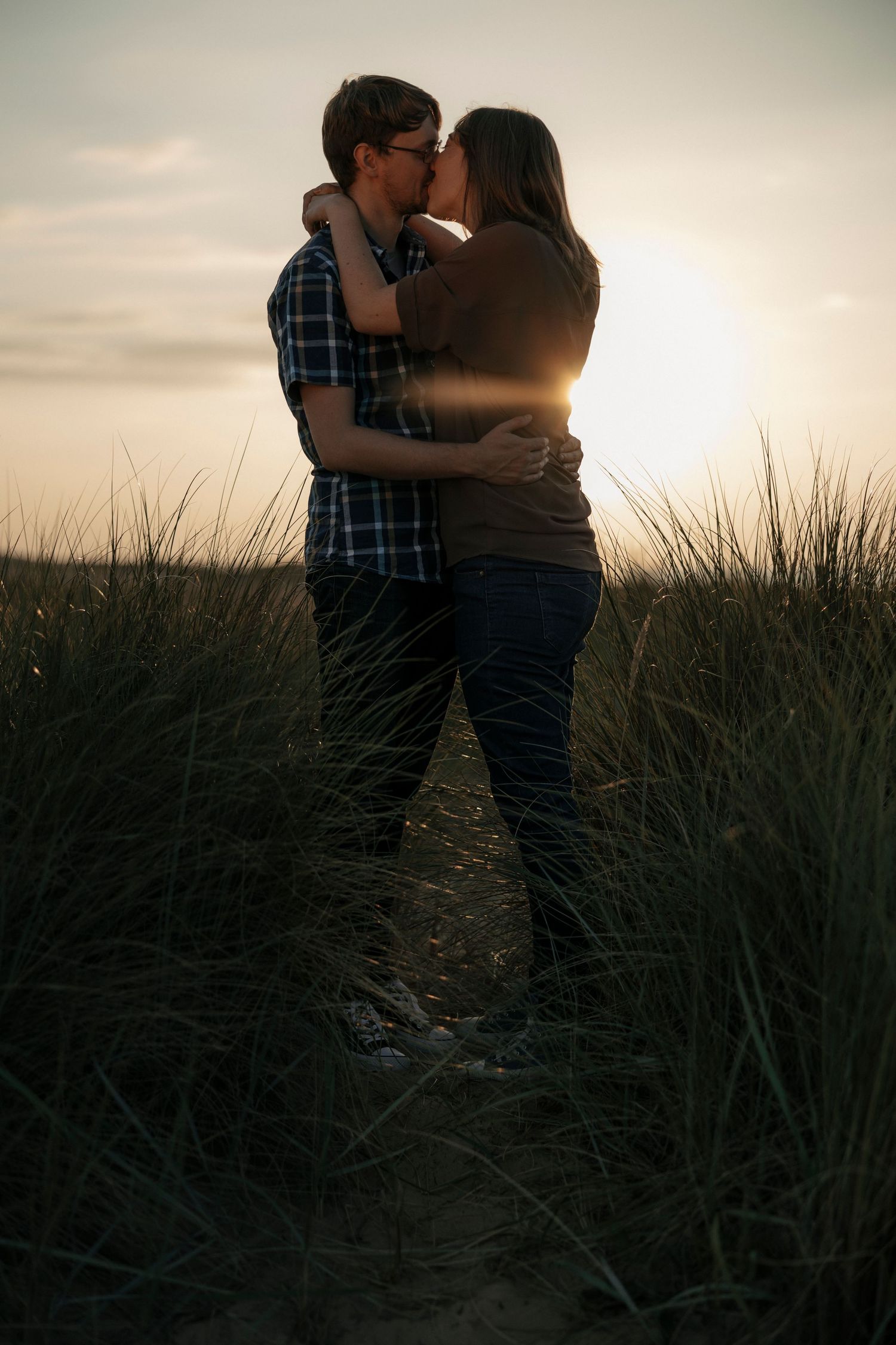 Romantic couple kissing silhouetted against stunning sunset in tall grass dunes.