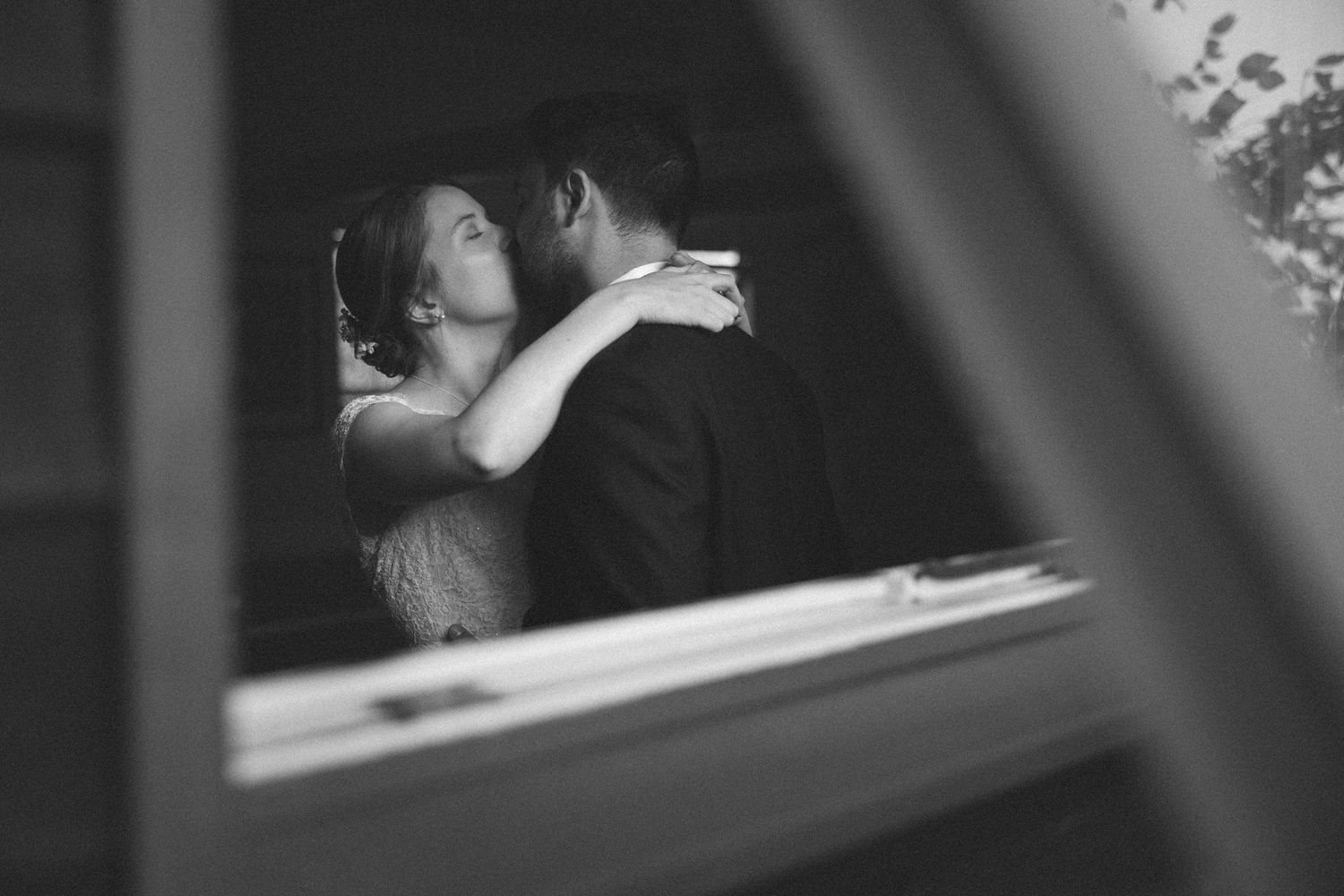 Black and white photo of a couple sharing a romantic embrace, viewed through a window frame.