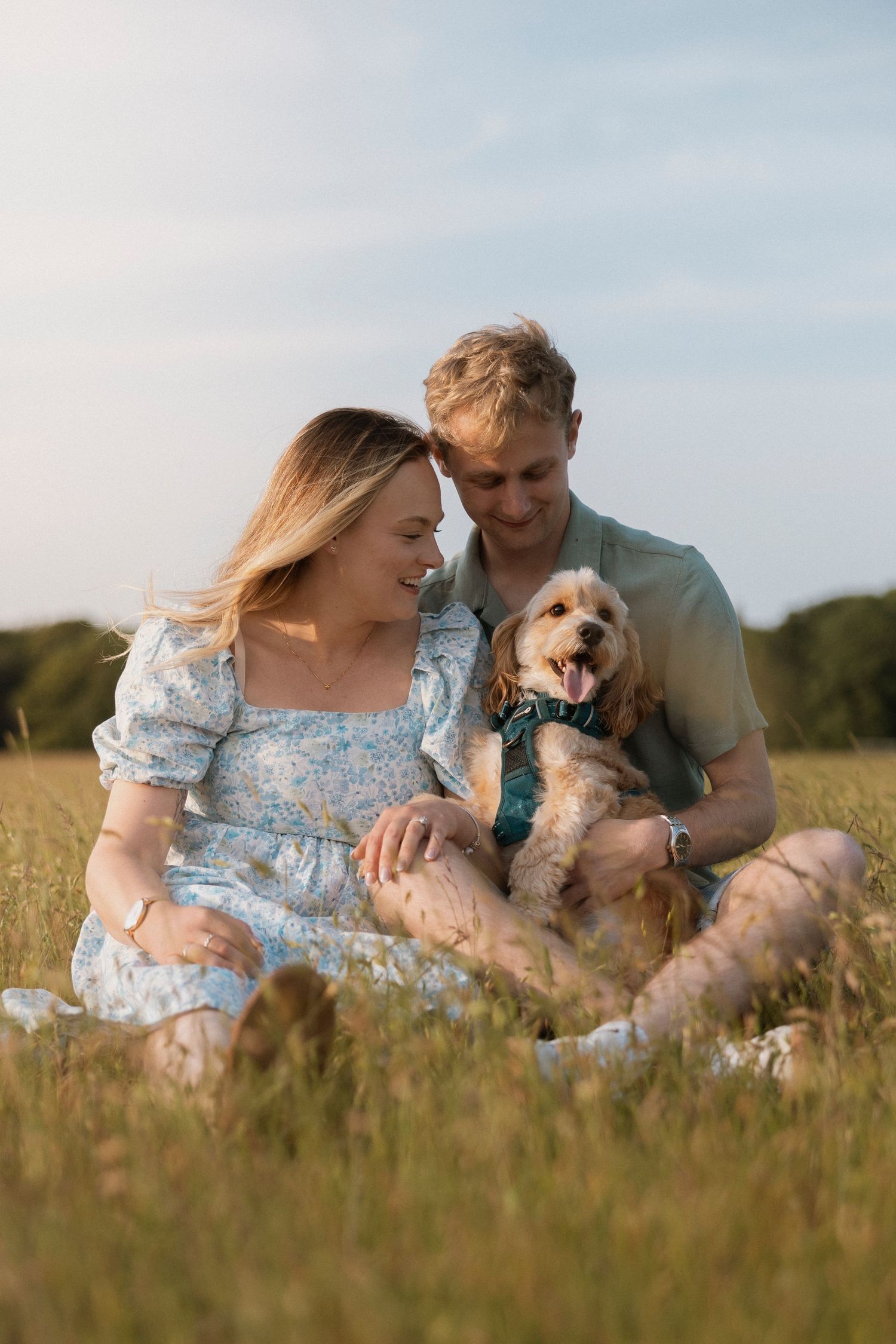 A smiling couple sits in a sunny green field with their happy golden dog, enjoying a relaxed outdoor moment together.