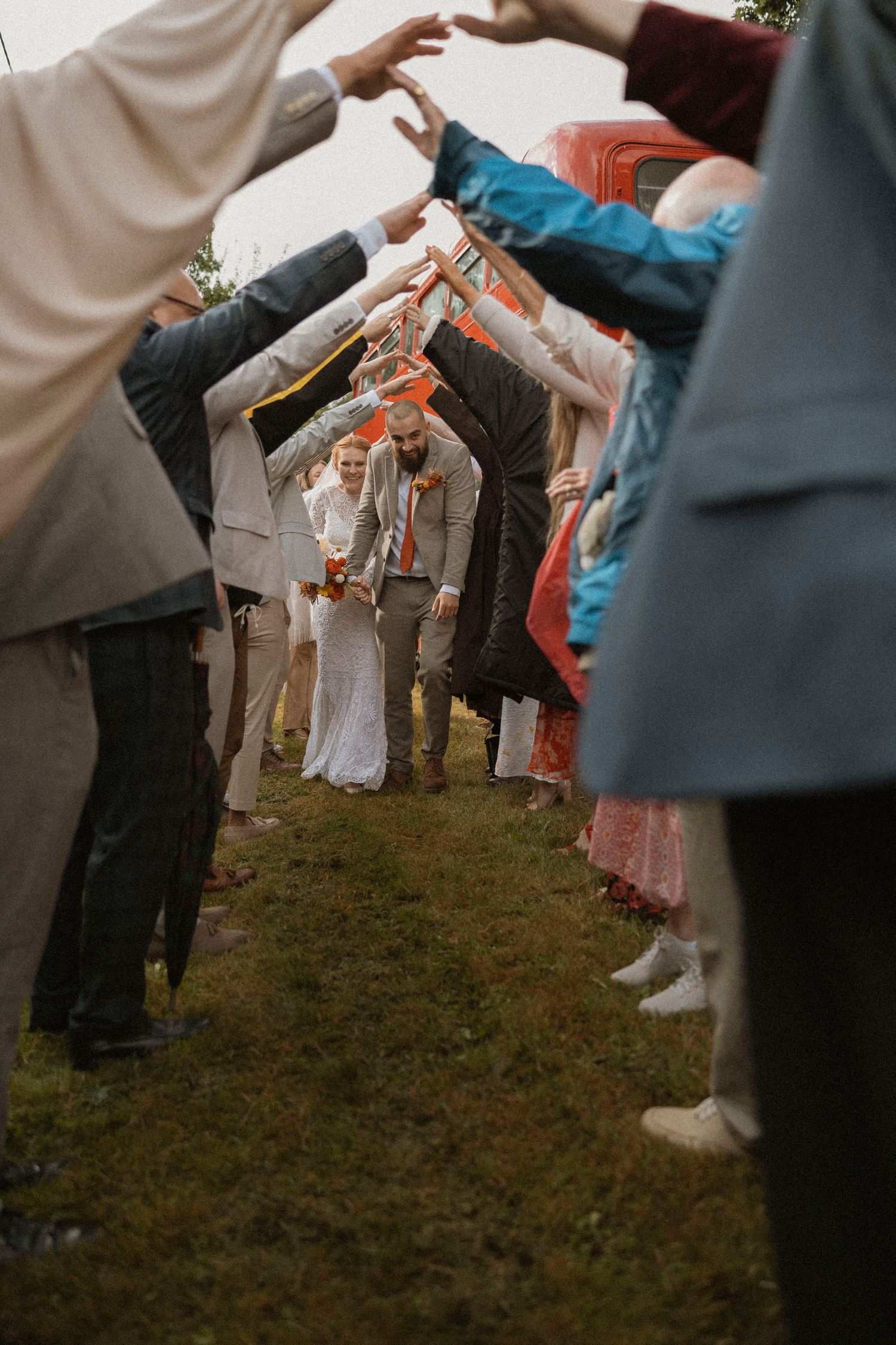 Wedding couple walks through tunnel of raised hands formed by guests outdoors on grass.