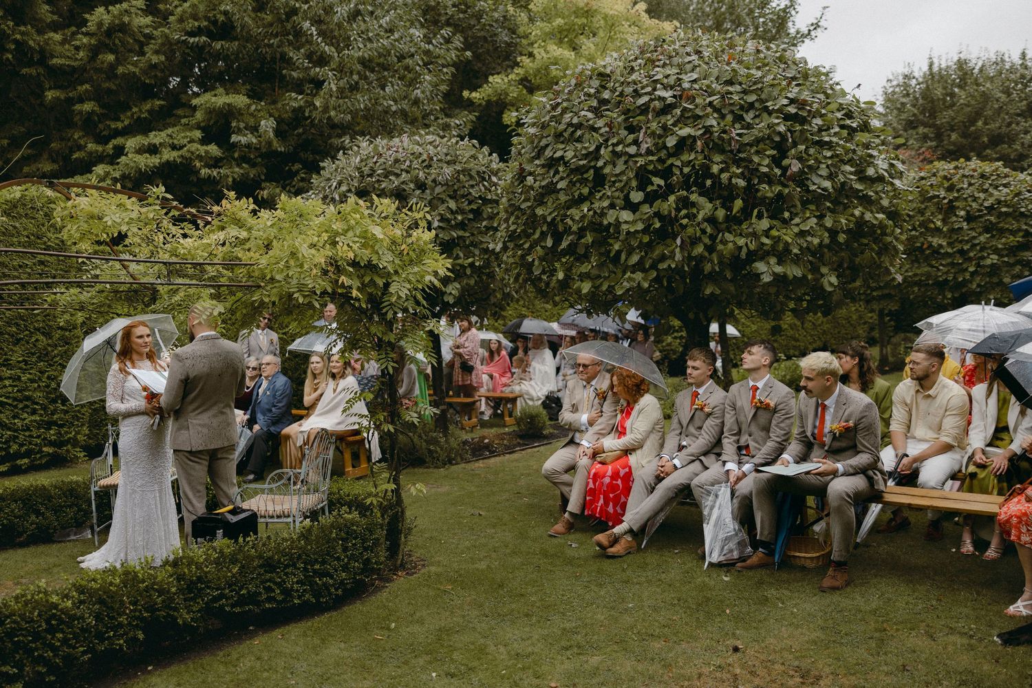 Outdoor garden wedding ceremony with guests holding umbrellas seated on benches, bride and groom at altar surrounded by lush greenery.