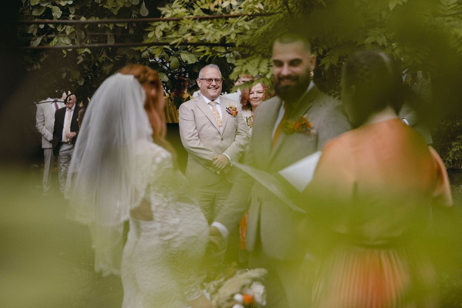 Outdoor wedding ceremony with smiling guests, groom in gray suit, bride in white gown and veil, surrounded by lush greenery.