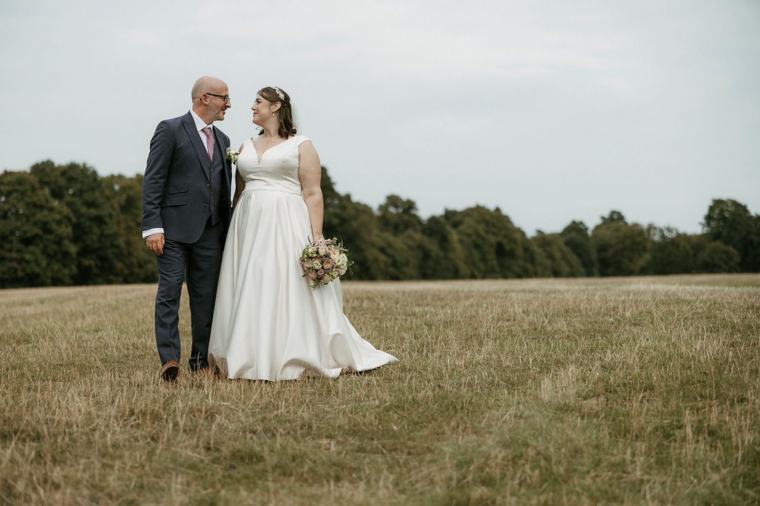 Bride and groom standing in an open field, sharing a romantic moment, bride holding a bouquet in a white gown.