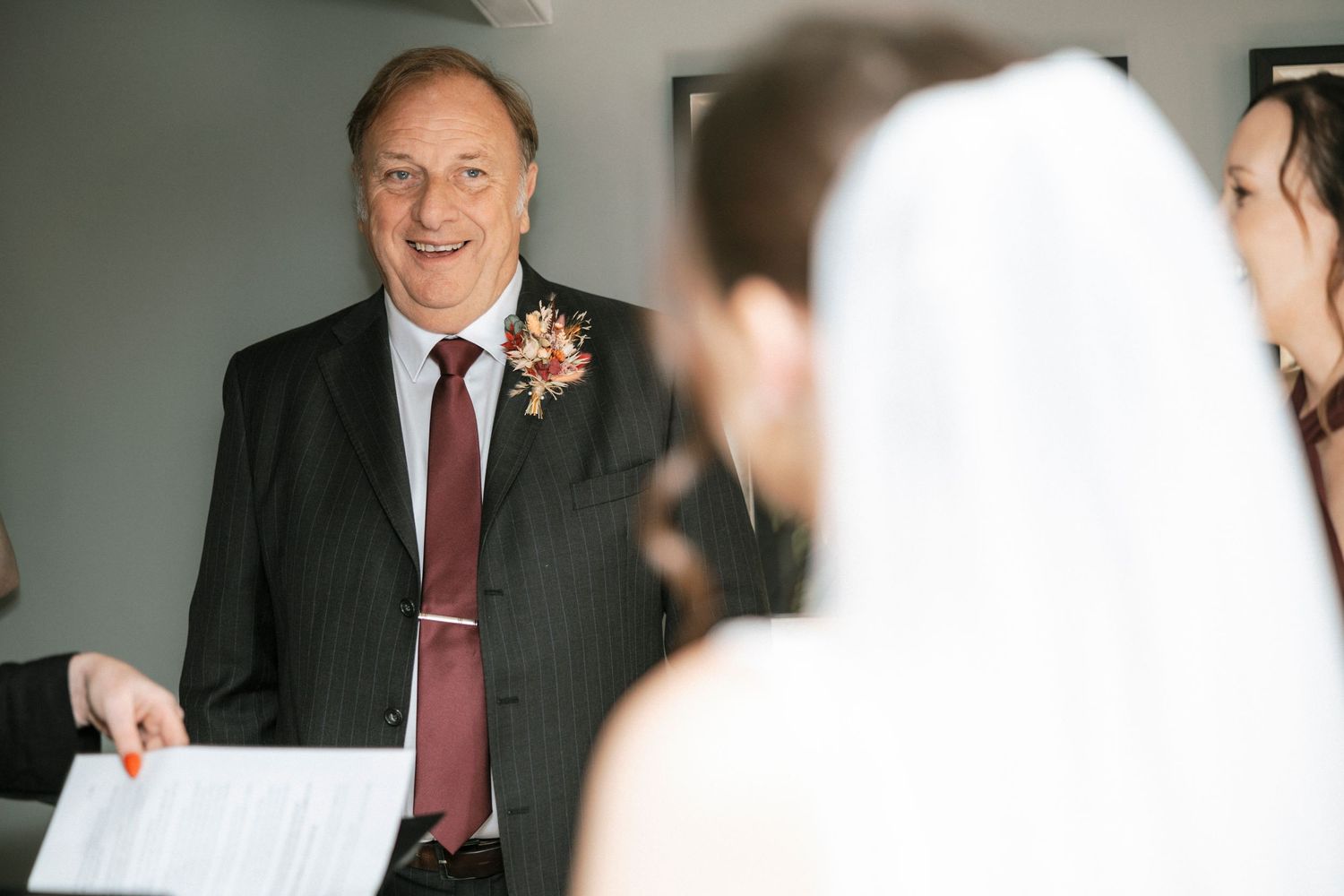 Smiling officiant in pinstripe suit holds ceremony papers while addressing bride and groom at wedding.
