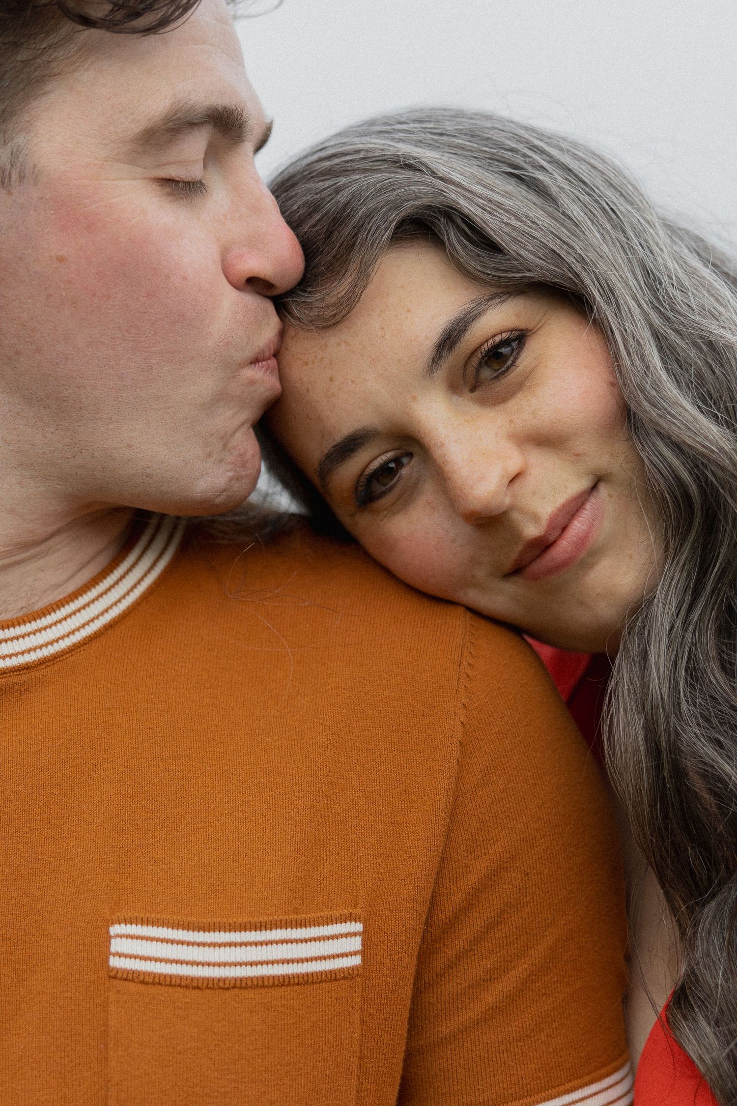 A man in an orange shirt kisses a woman with gray hair on the head as she smiles and leans against him.