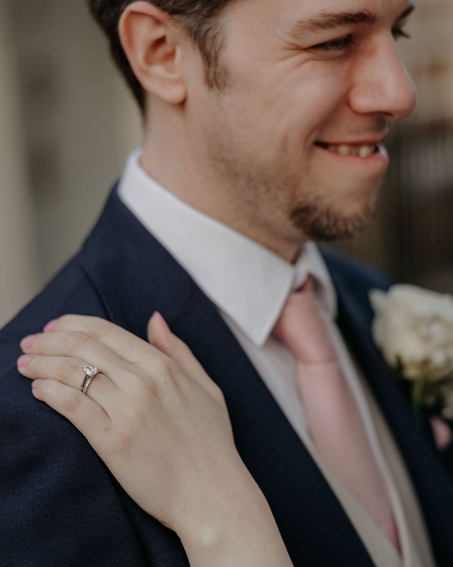 Groom in navy suit and blush pink tie with bride's hand showing diamond engagement ring on his shoulder.