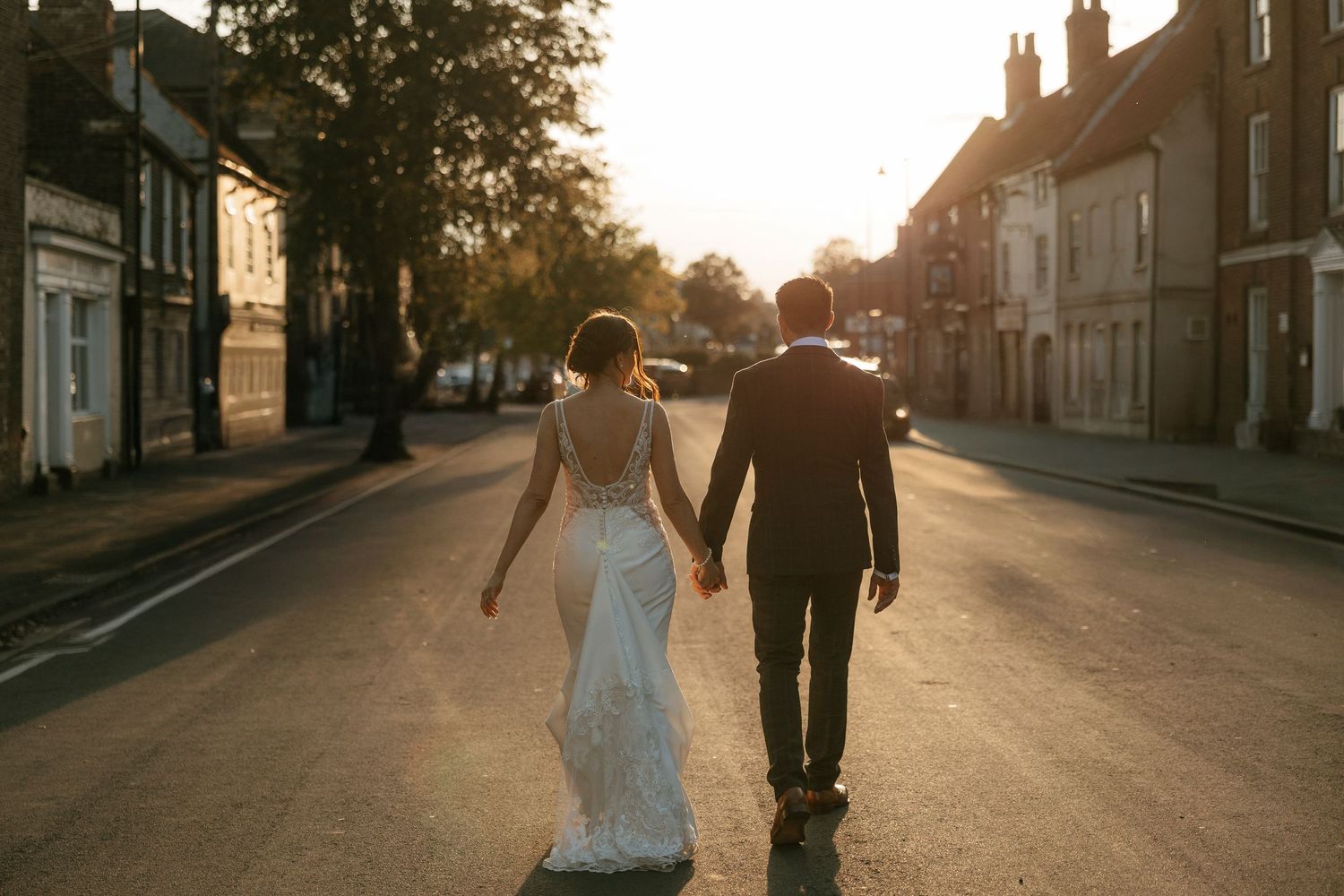 Bride and groom walk hand in hand down a sunlit street at golden hour in a charming English town.