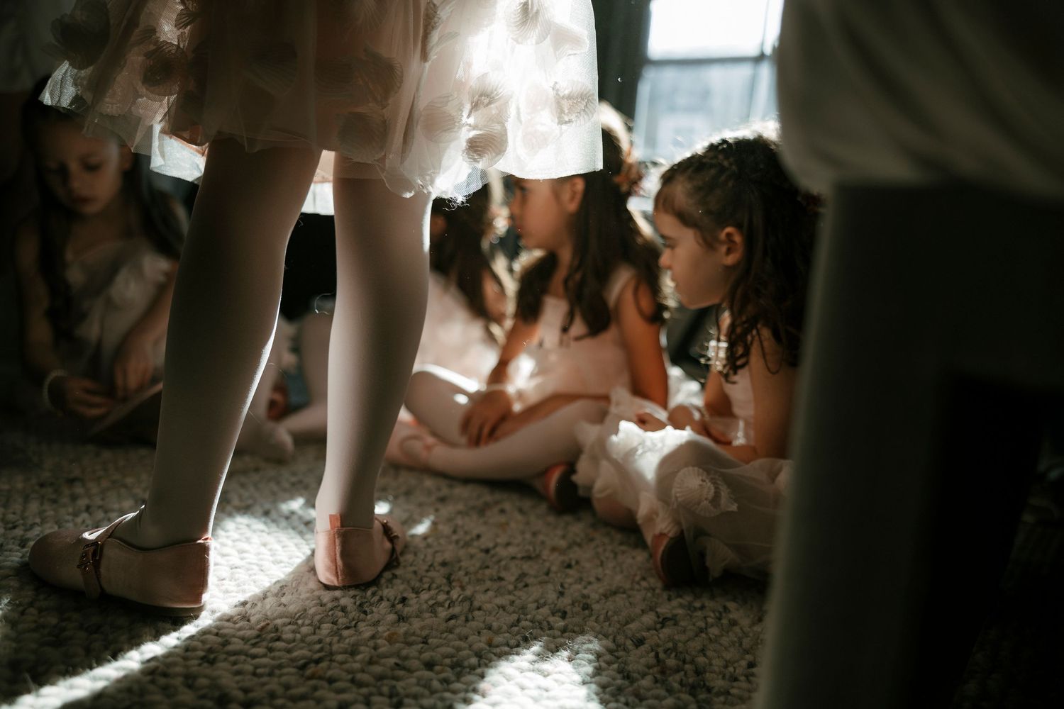 Young flower girls in white dresses sitting on carpet in soft natural light during a wedding preparation.