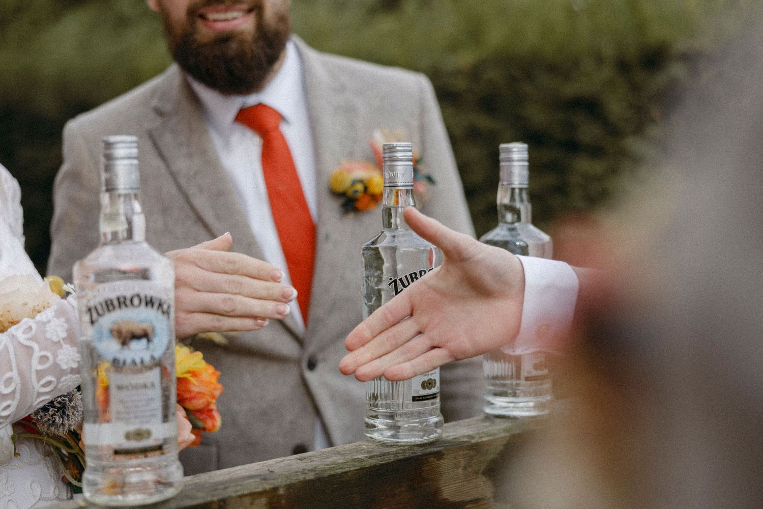 Groom in gray suit with orange tie holding a tray of Zubrowka vodka bottles at a wedding reception.