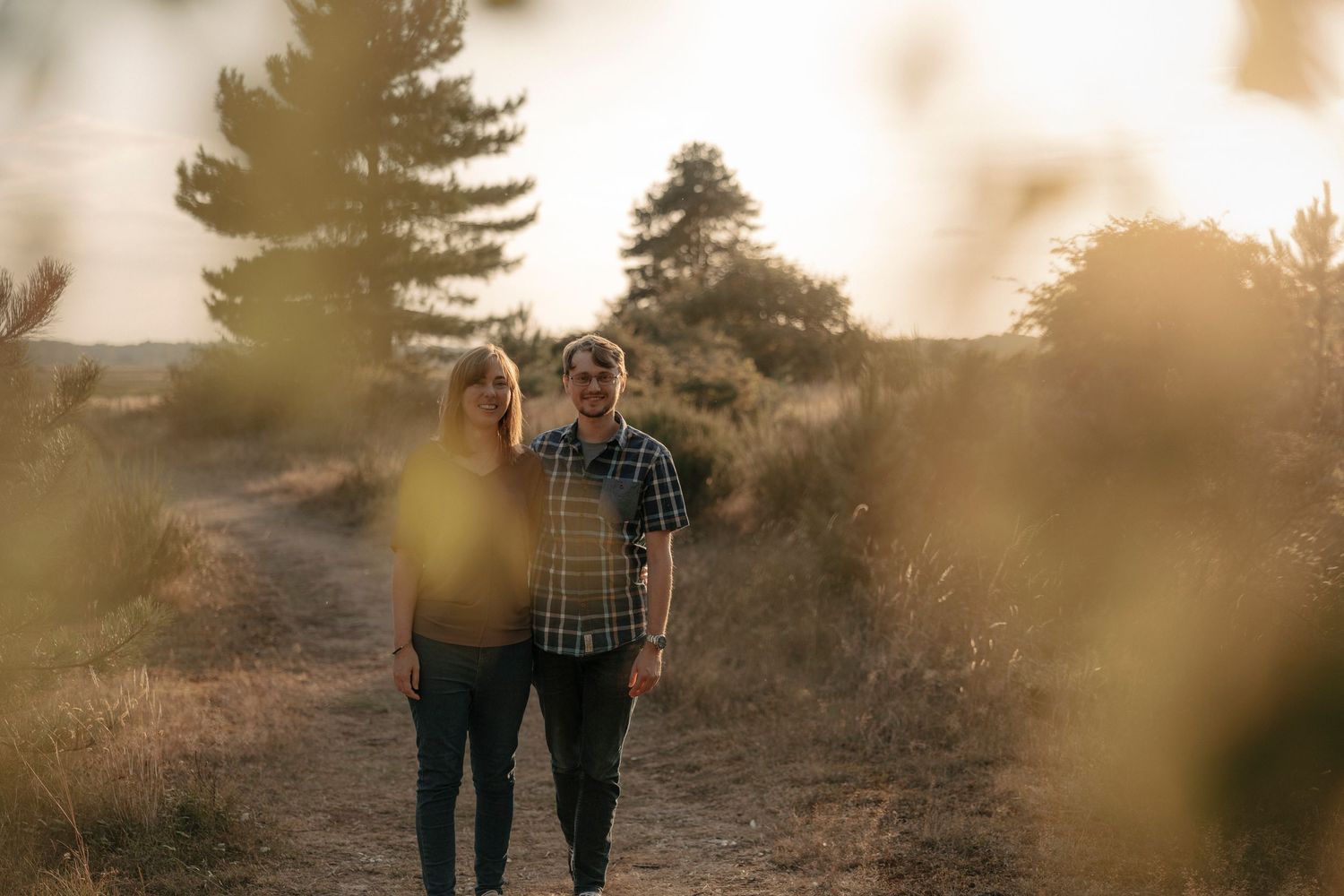 Couple walking on a sunlit dirt path surrounded by golden trees and shrubs during sunset.