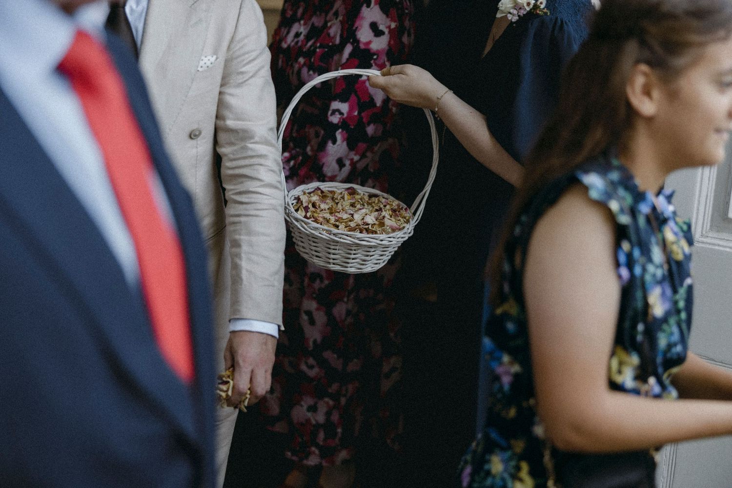 A child holds a wicker basket of petals at a wedding, surrounded by guests in formal attire.