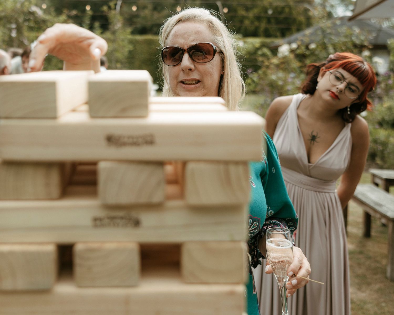 Two women play giant Jenga at an outdoor wedding reception, one wearing sunglasses and a teal wrap.