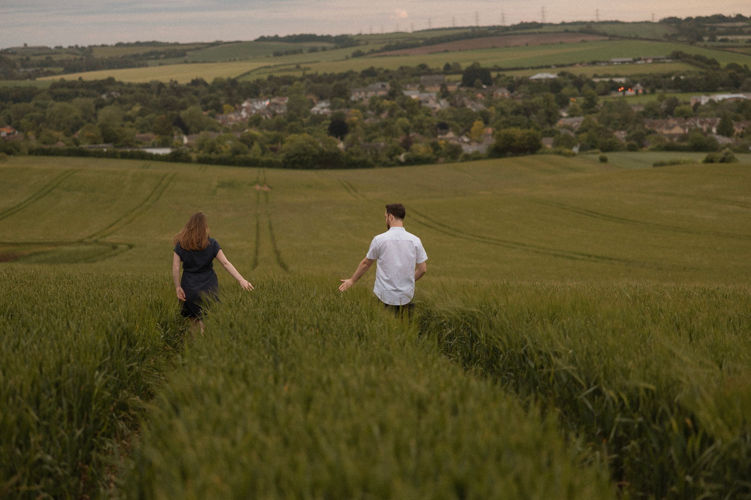 Two people walking down a grassy hillside path overlooking a scenic countryside valley.