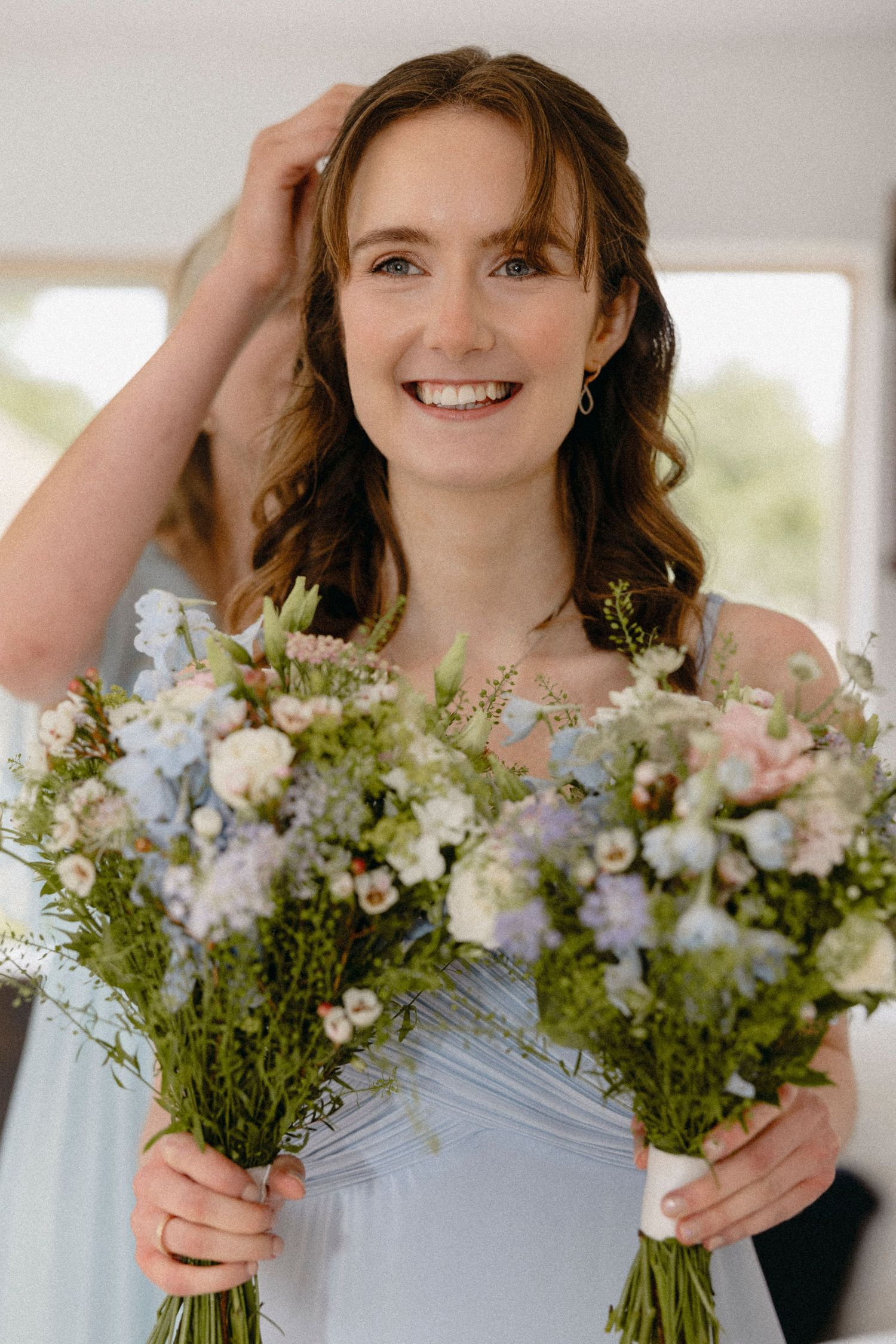Smiling bridesmaid in light blue dress holding two pastel wildflower bouquets with blue, pink, and white blooms.