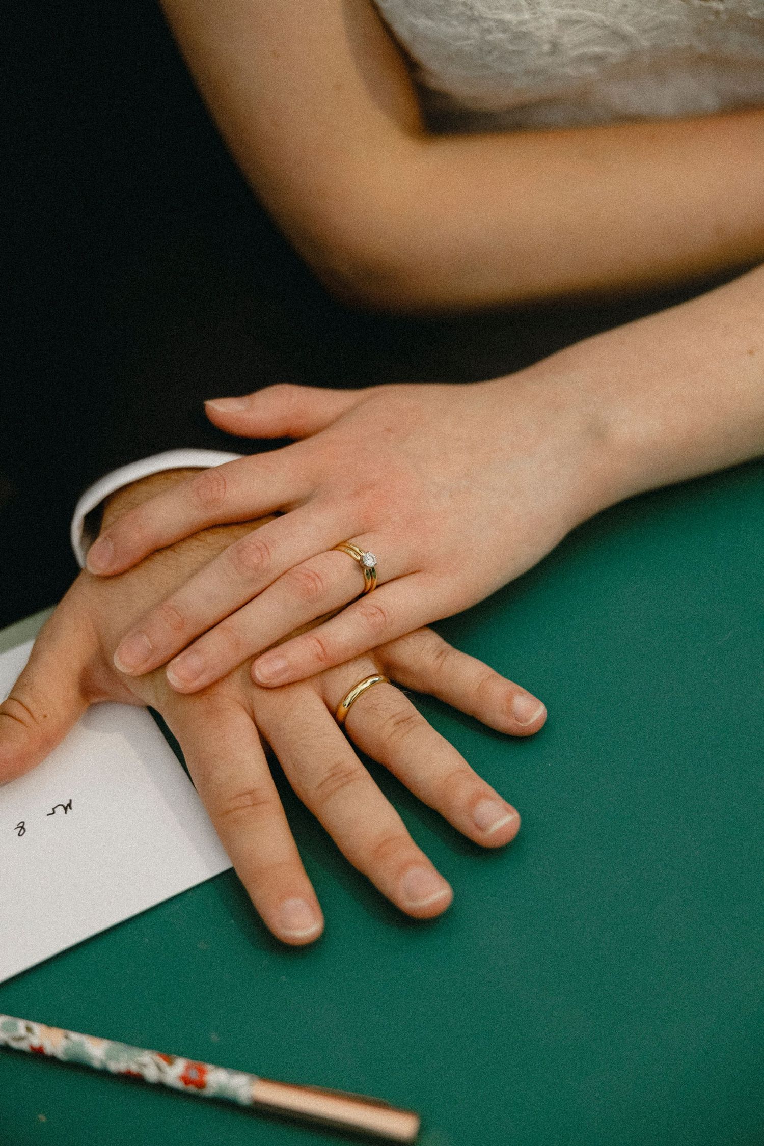Newlyweds hands with gold wedding rings resting on a green table beside a pen and paper.