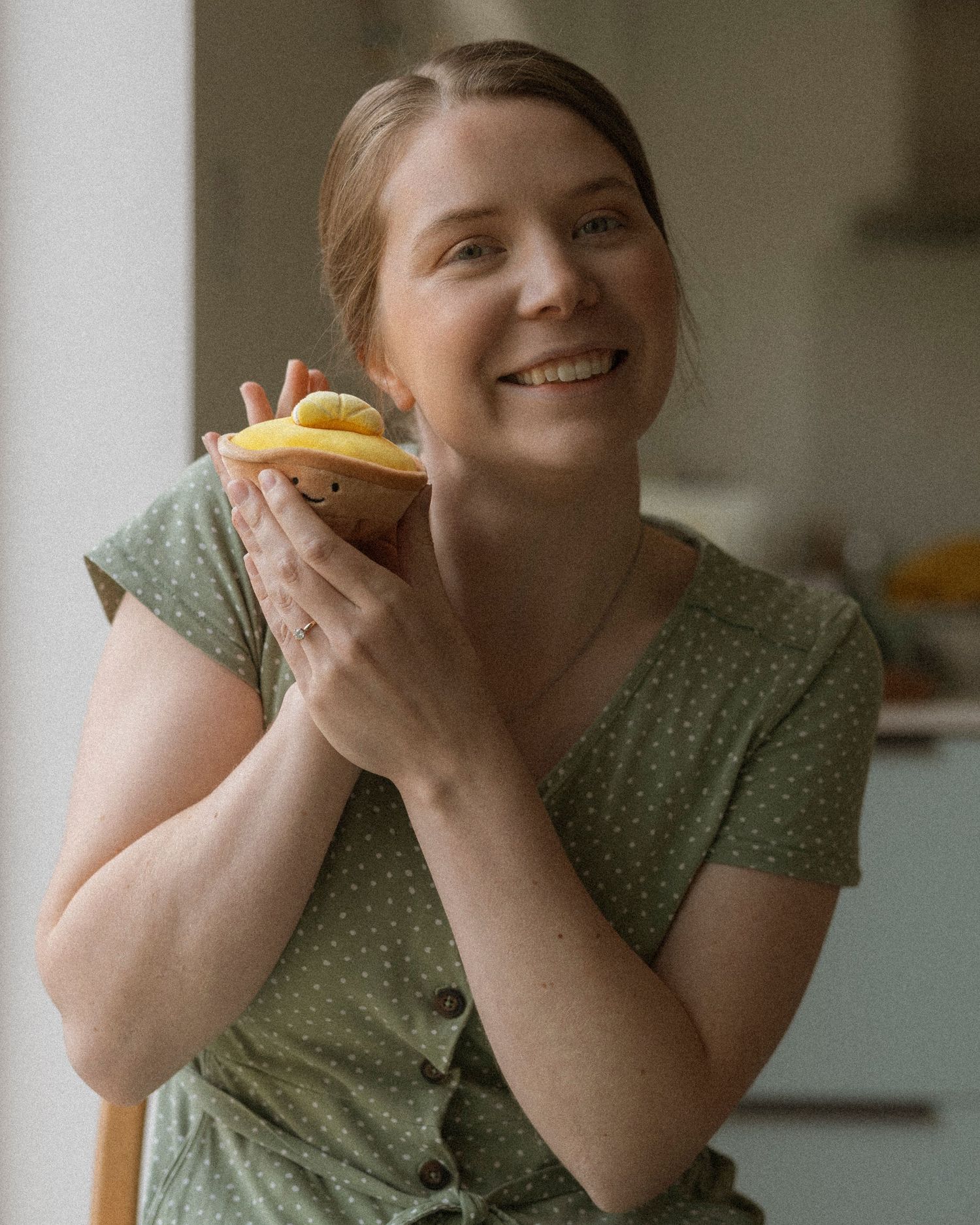 Smiling woman in green polka dot dress holding a colorful cupcake in a bright kitchen setting.