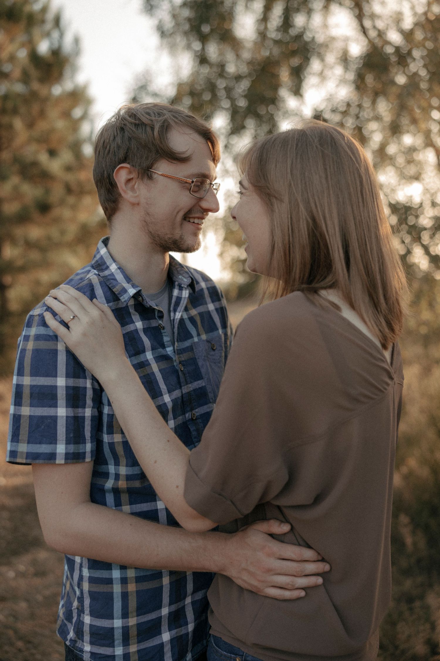A smiling couple in a warm embrace, gazing into each other's eyes in a sunlit autumn forest setting.