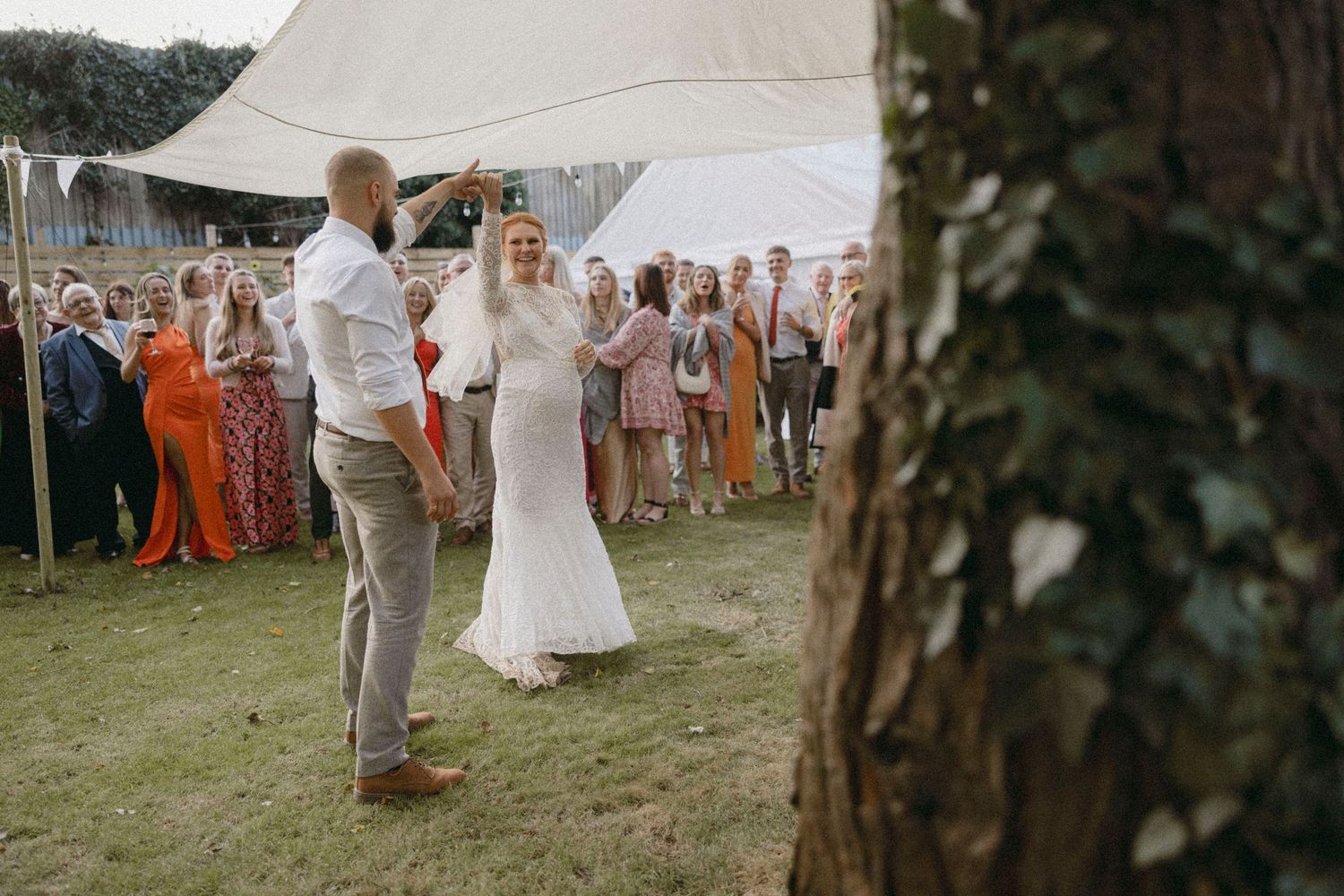 Bride and groom share first dance outdoors at elegant garden wedding reception under white tent with guests watching.