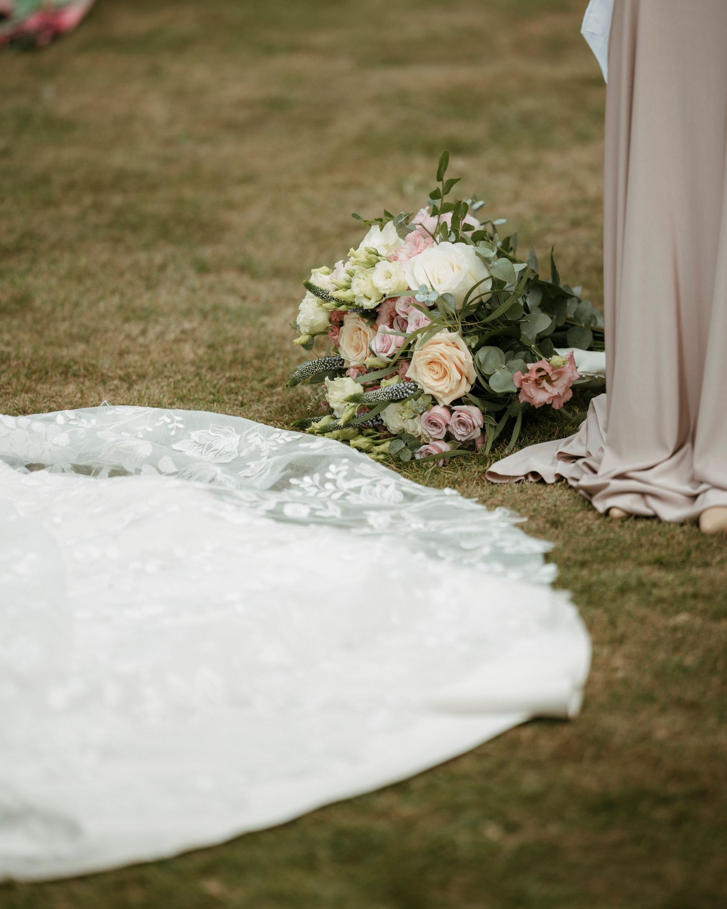 Wedding dress train and bridal bouquet of pink and white roses resting on grass outdoors.
