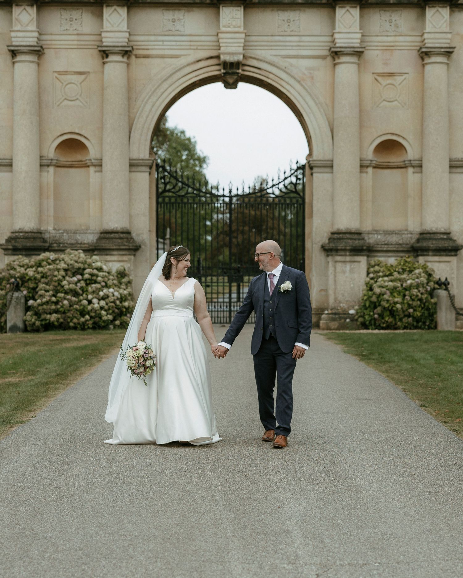 Bride and groom hold hands walking toward ornate stone arch gate at elegant wedding venue.