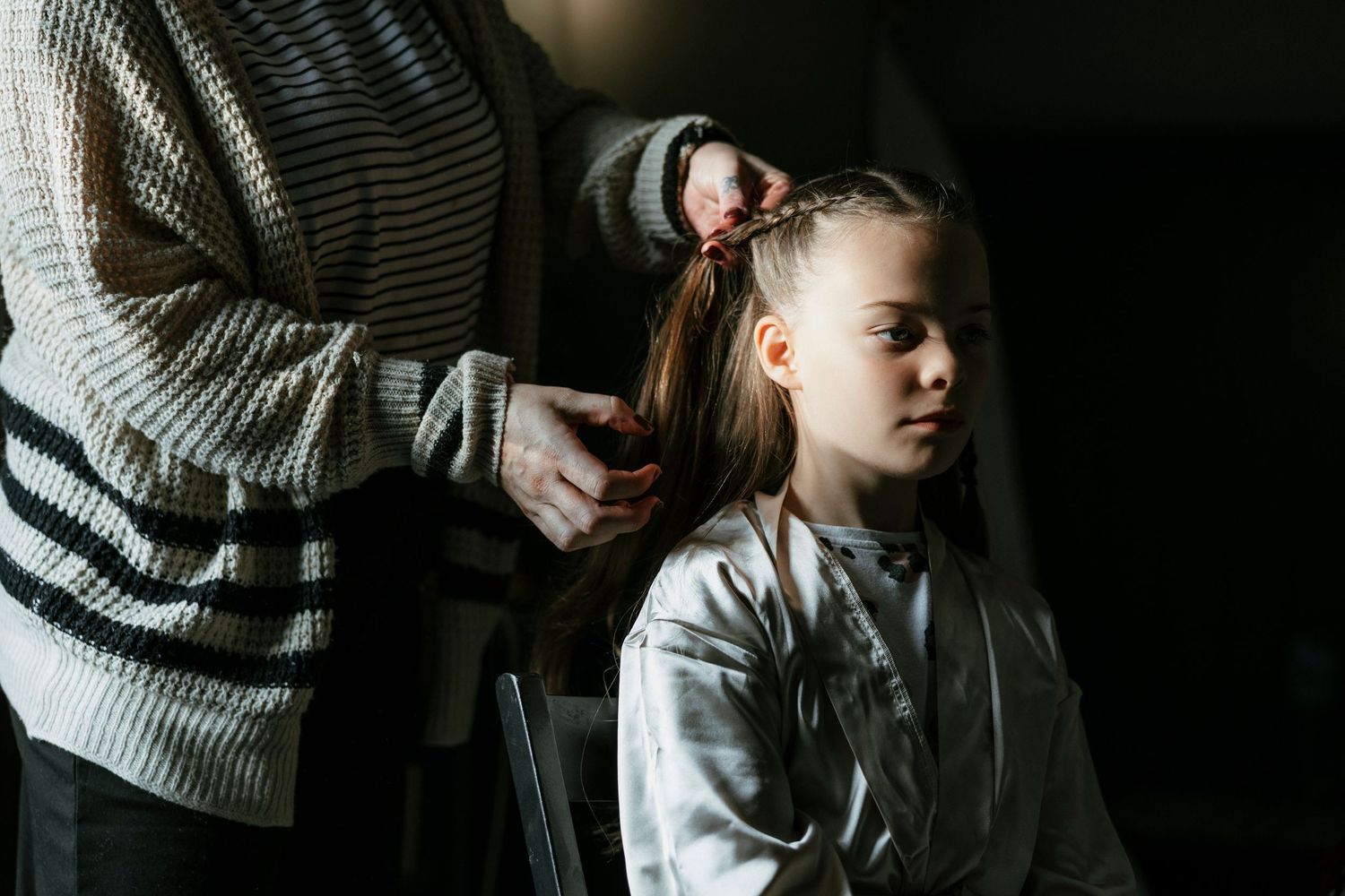 Adult braiding young girl's hair in a dimly lit room, child sitting calmly in chair wearing gray jacket.