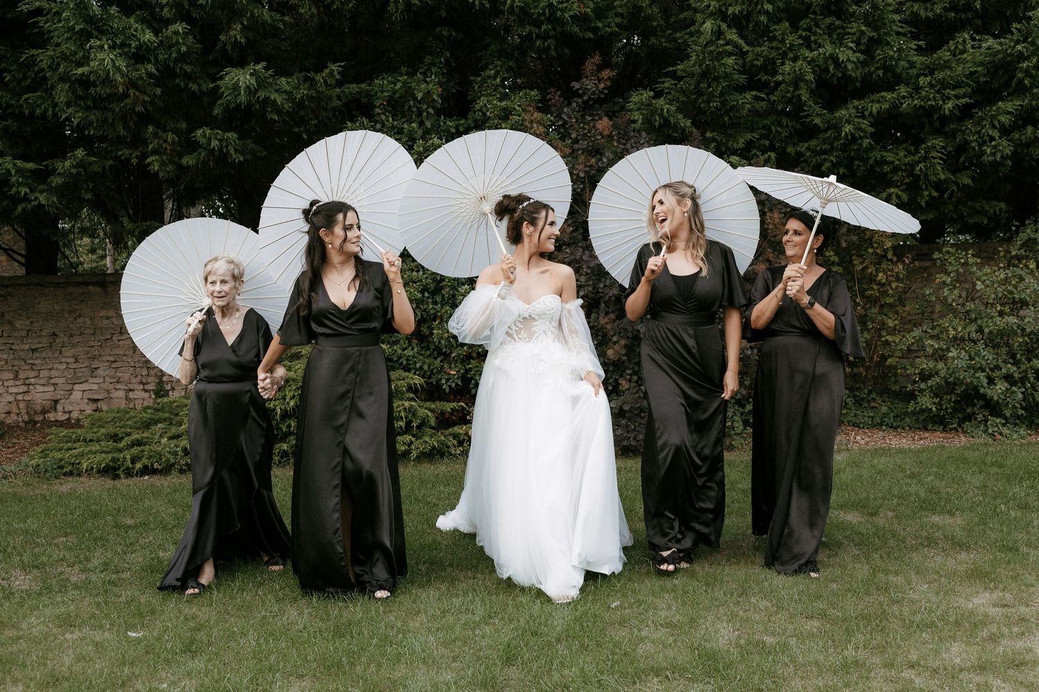 Bride in white gown with four bridesmaids in black dresses holding white parasols outdoors at a wedding.