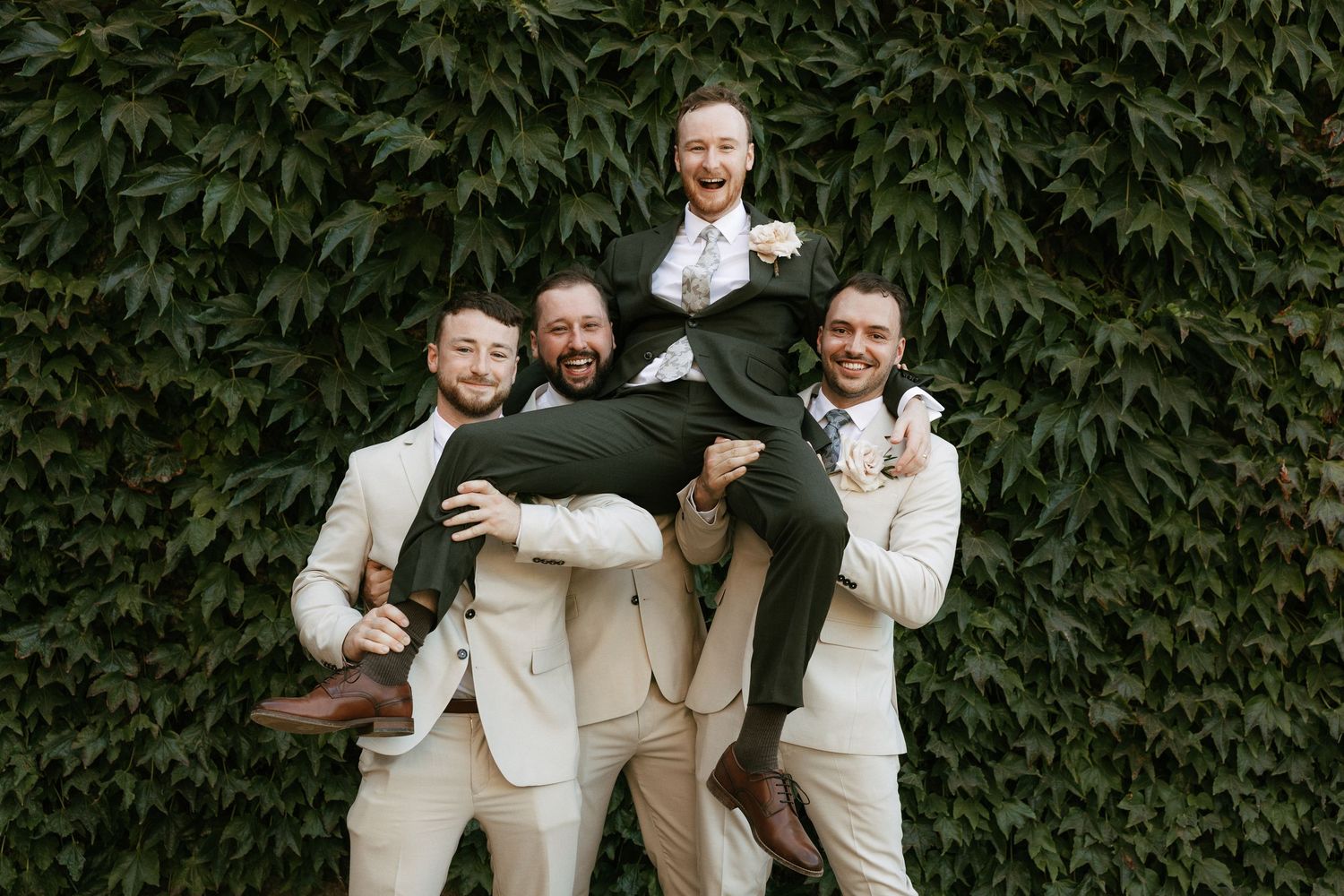 Groomsmen in cream suits joyfully lift the groom in a dark suit against a lush ivy wall.