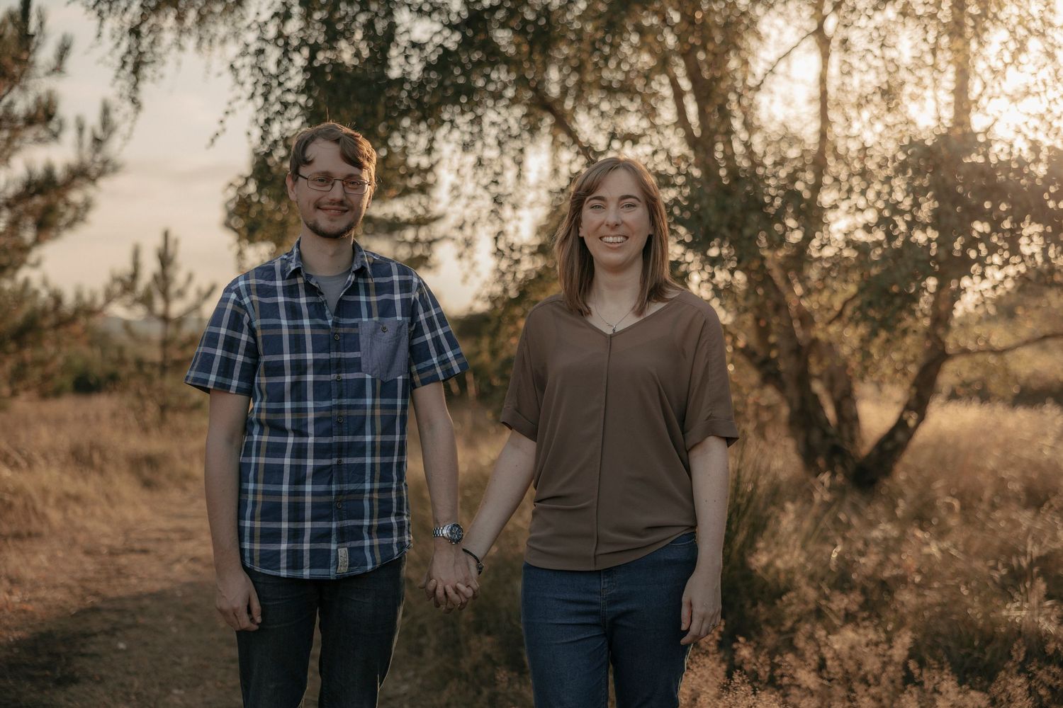 Couple holding hands smiling in golden sunset light among trees and dry grass outdoors.