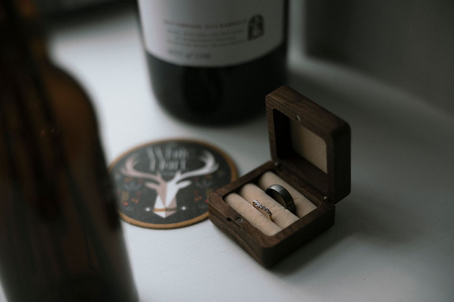 Small wooden box with rings sits next to wine bottle and coaster on table.