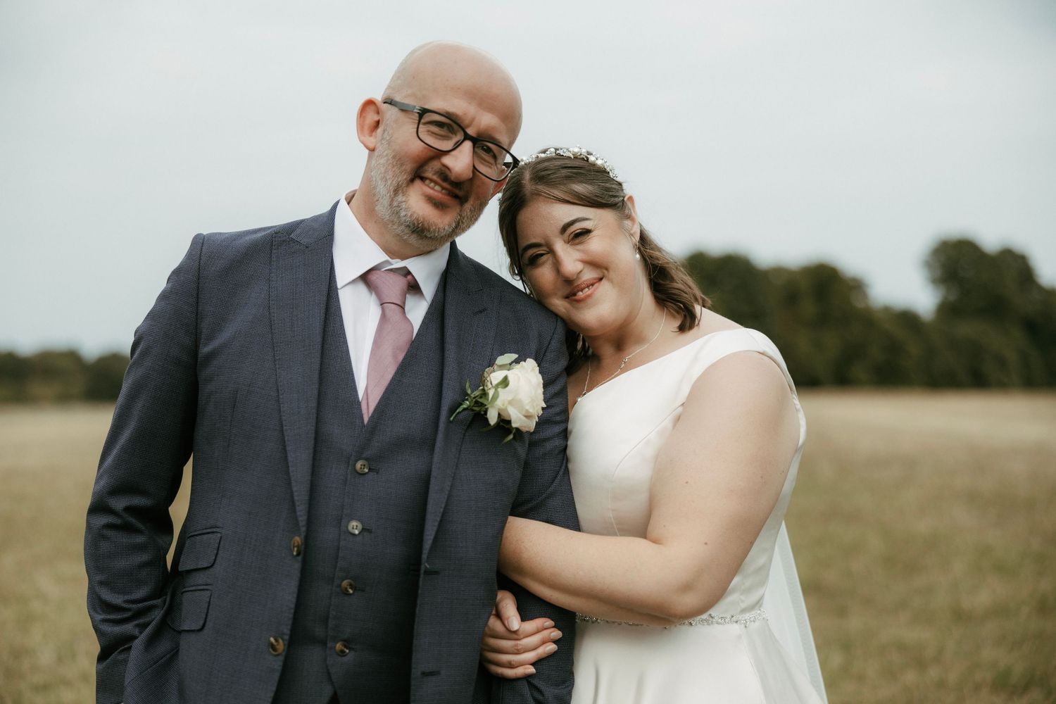 Bride and groom smiling together in a field on their wedding day, groom in navy suit, bride in white dress.