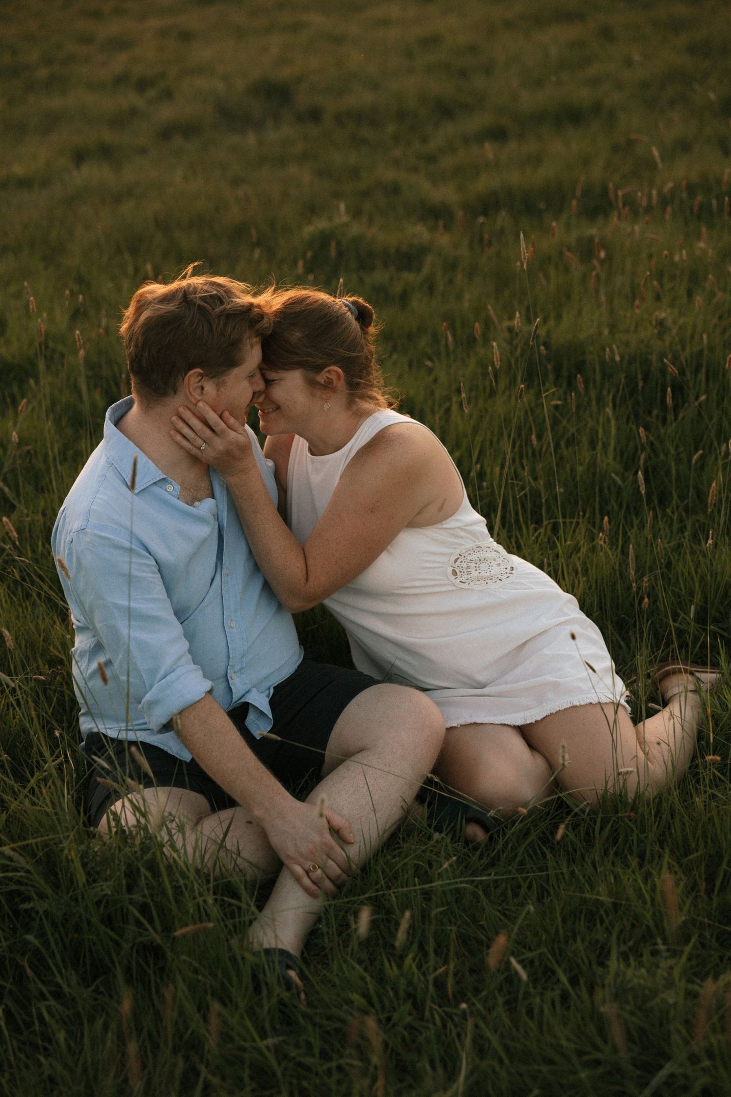 Couple sharing a romantic moment sitting in a lush green field at golden hour, woman in white dress touching mans face.