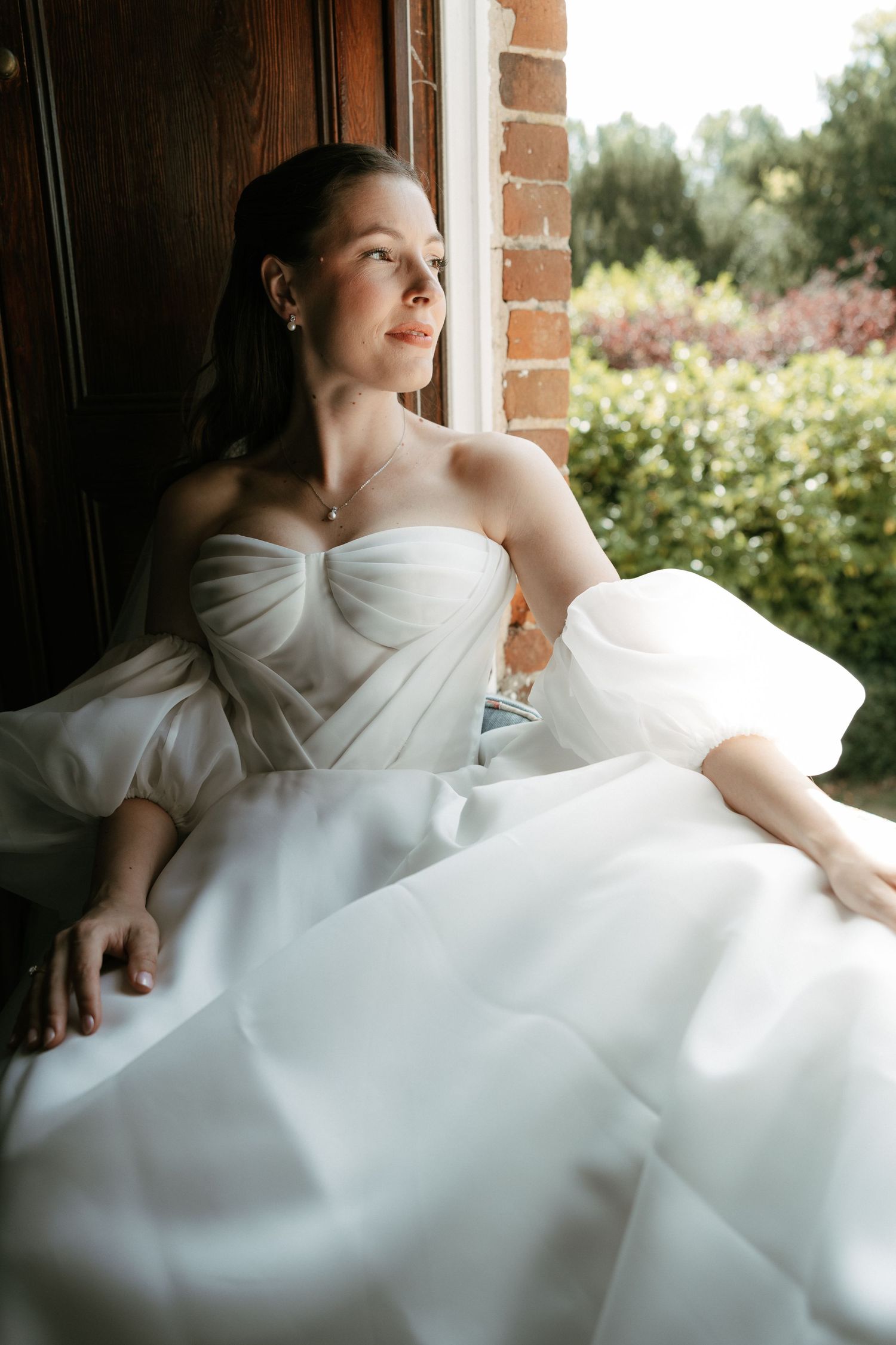 Bride in strapless white ballgown sitting by window with lush garden view, looking away dreamily.