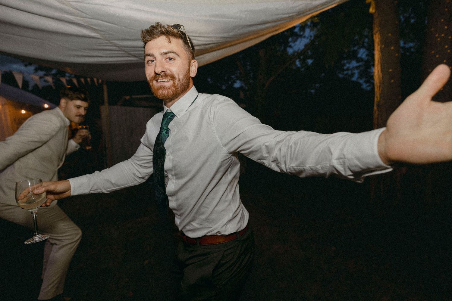 Man in green tie dancing with arms outstretched at an outdoor evening wedding reception celebration.