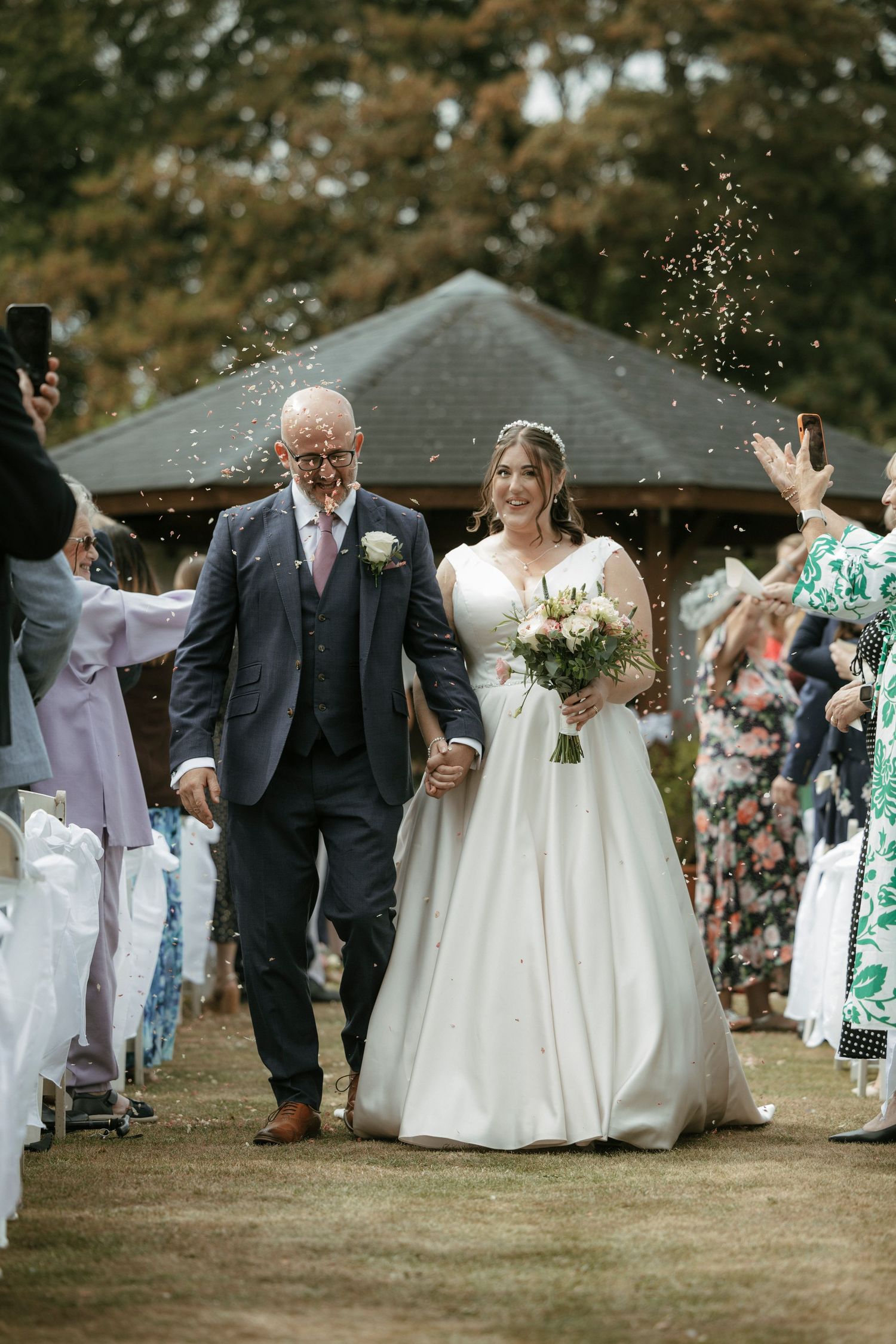 Newlyweds walking hand in hand through confetti shower at outdoor wedding ceremony with guests celebrating.