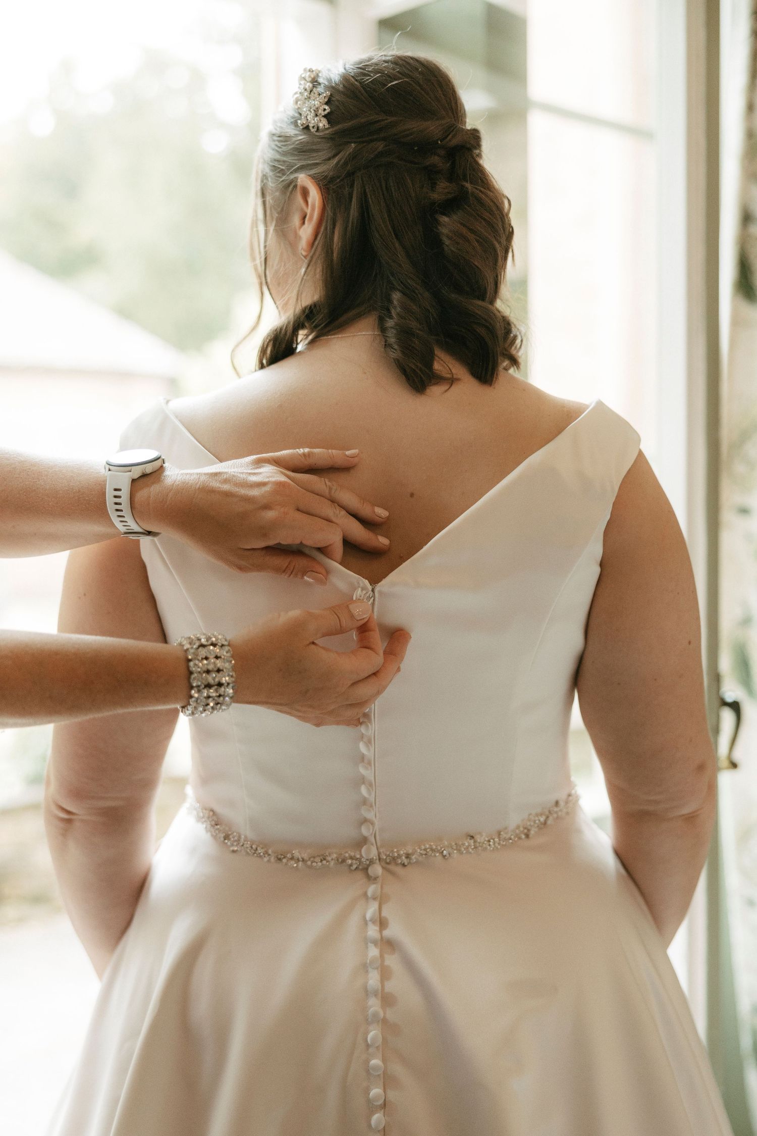 Bride's back view showing elegant white wedding dress with button detail and beaded belt being fastened by helper.