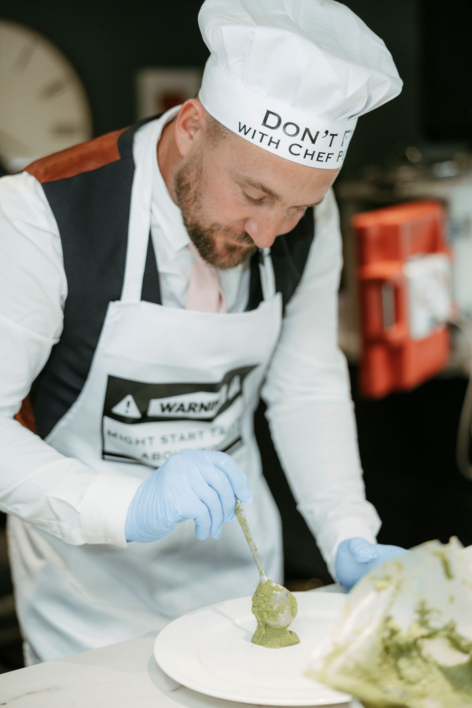Male chef in white apron and blue gloves carefully plates a green sauce on a white dish in a kitchen.